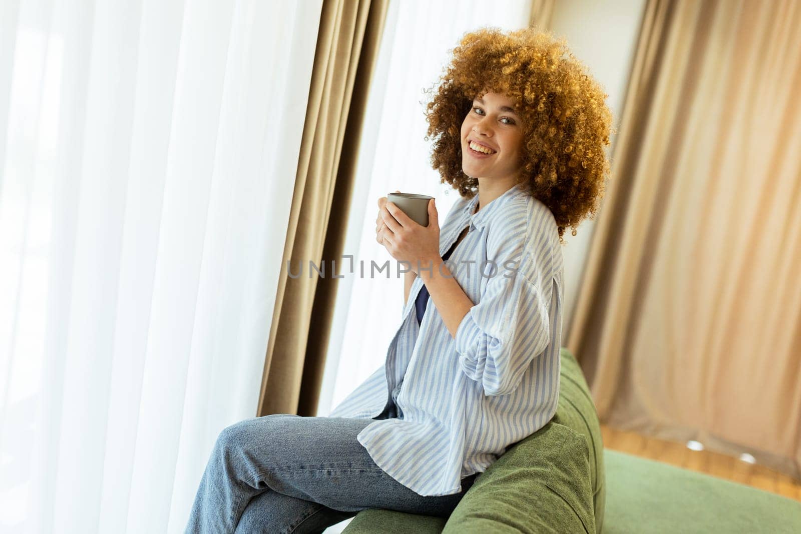 A young woman with curly hair is seated on a soft sofa, holding a warm drink and looking cheerfully towards the camera, with sunlight streaming through the window