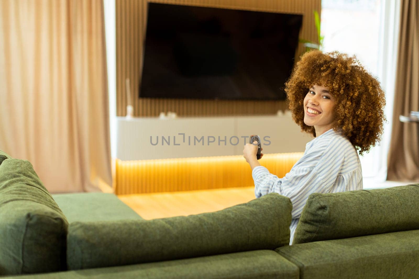 A woman with curly hair beams with joy as she sits on a soft couch, ready to enjoy her favorite show while holding the remote control in a modern living space
