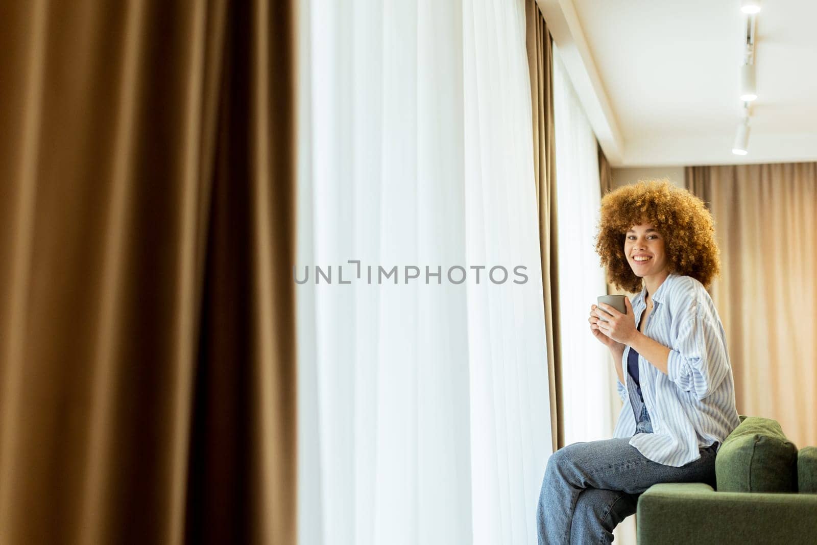 A young woman with curly hair is seated on a soft sofa, holding a warm drink and looking cheerfully towards the camera, with sunlight streaming through the window
