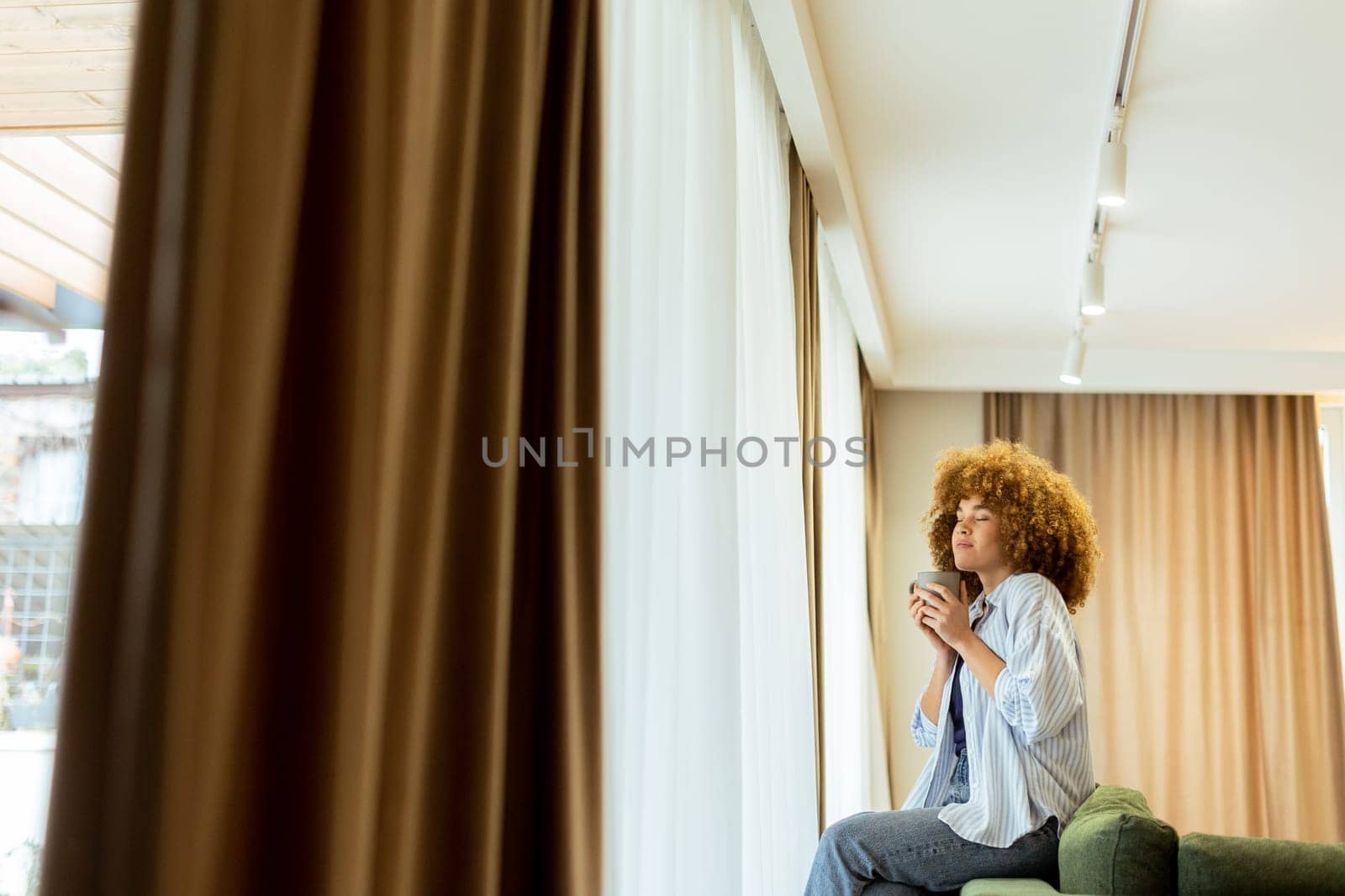 A young woman with curly hair is seated on a soft sofa, holding a warm drink and looking cheerfully towards the camera, with sunlight streaming through the window