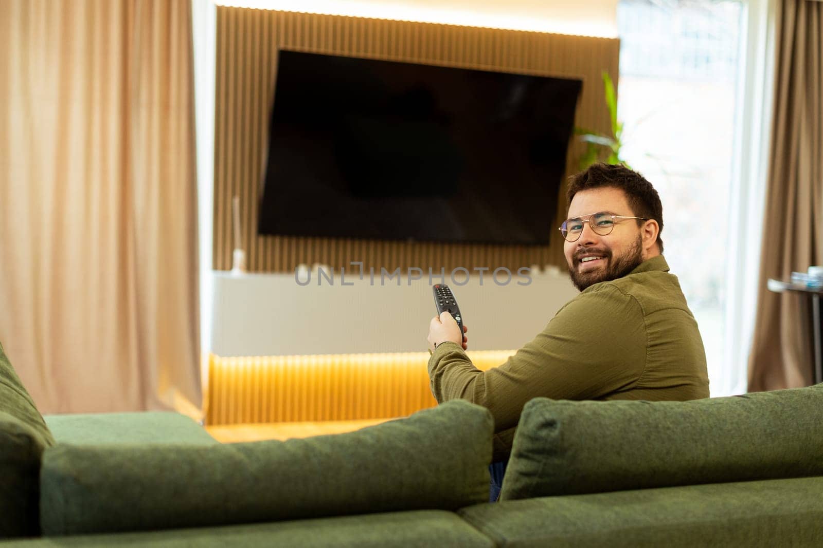 A man sits comfortably on a green couch, holding a remote, engaged in his favorite show. Soft lighting and modern decor create a serene ambiance perfect for unwinding.