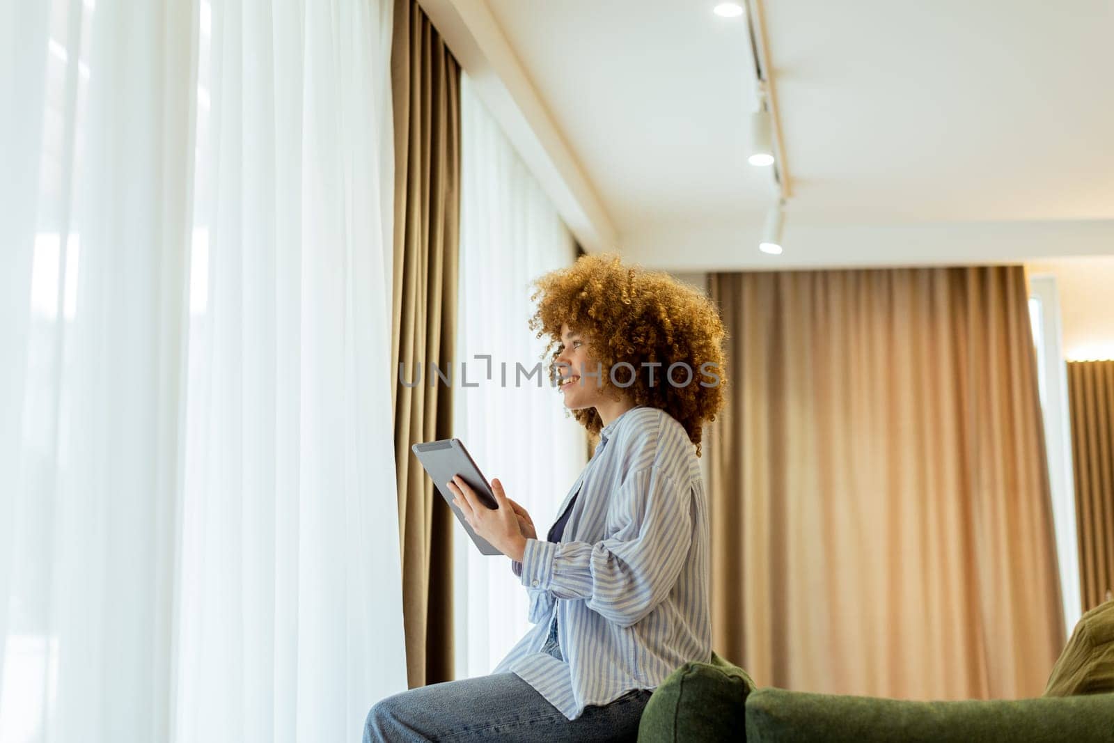 A woman with curly hair enjoys using her tablet, seated comfortably on a couch in a bright, inviting living space, surrounded by soft curtains and warm lighting