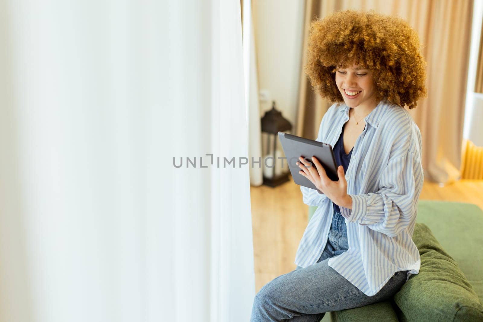 A woman with curly hair enjoys using her tablet, seated comfortably on a couch in a bright, inviting living space, surrounded by soft curtains and warm lighting