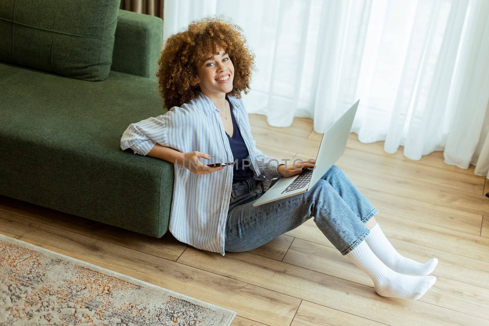 Seated comfortably on the floor, a young woman engages with her laptop while checking her smartphone. The cozy, sunlit living room creates a serene backdrop for her dual focus