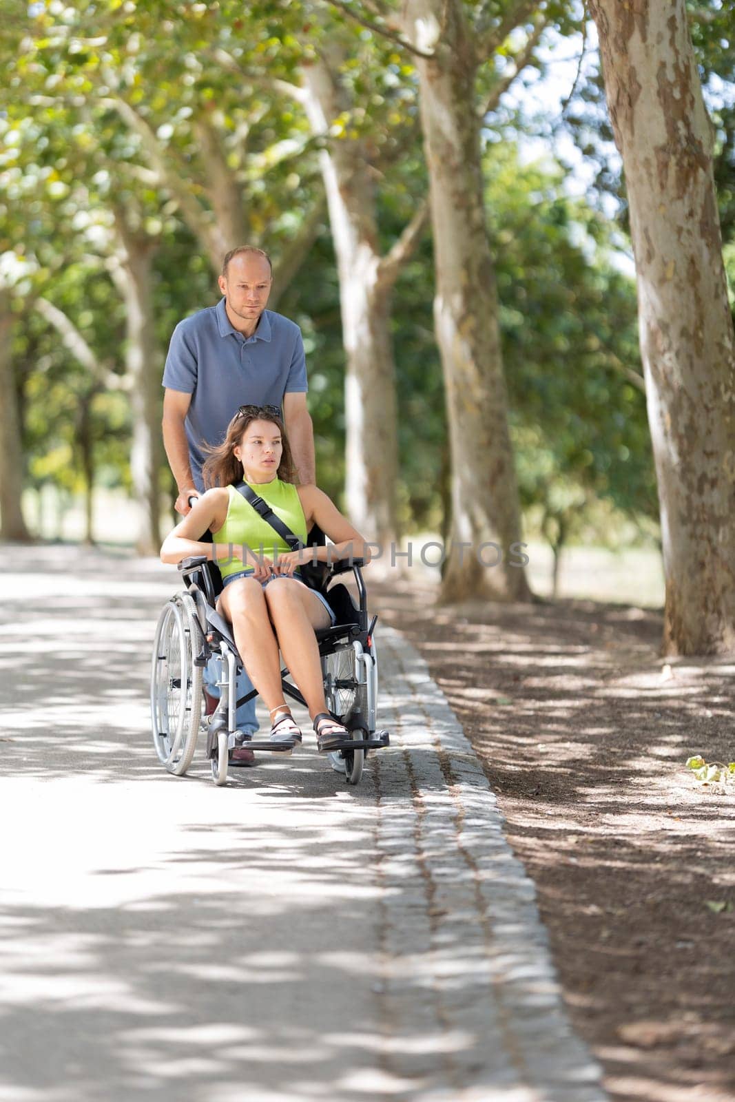 Nurse assisting patient in wheelchair along path in park on sunny day