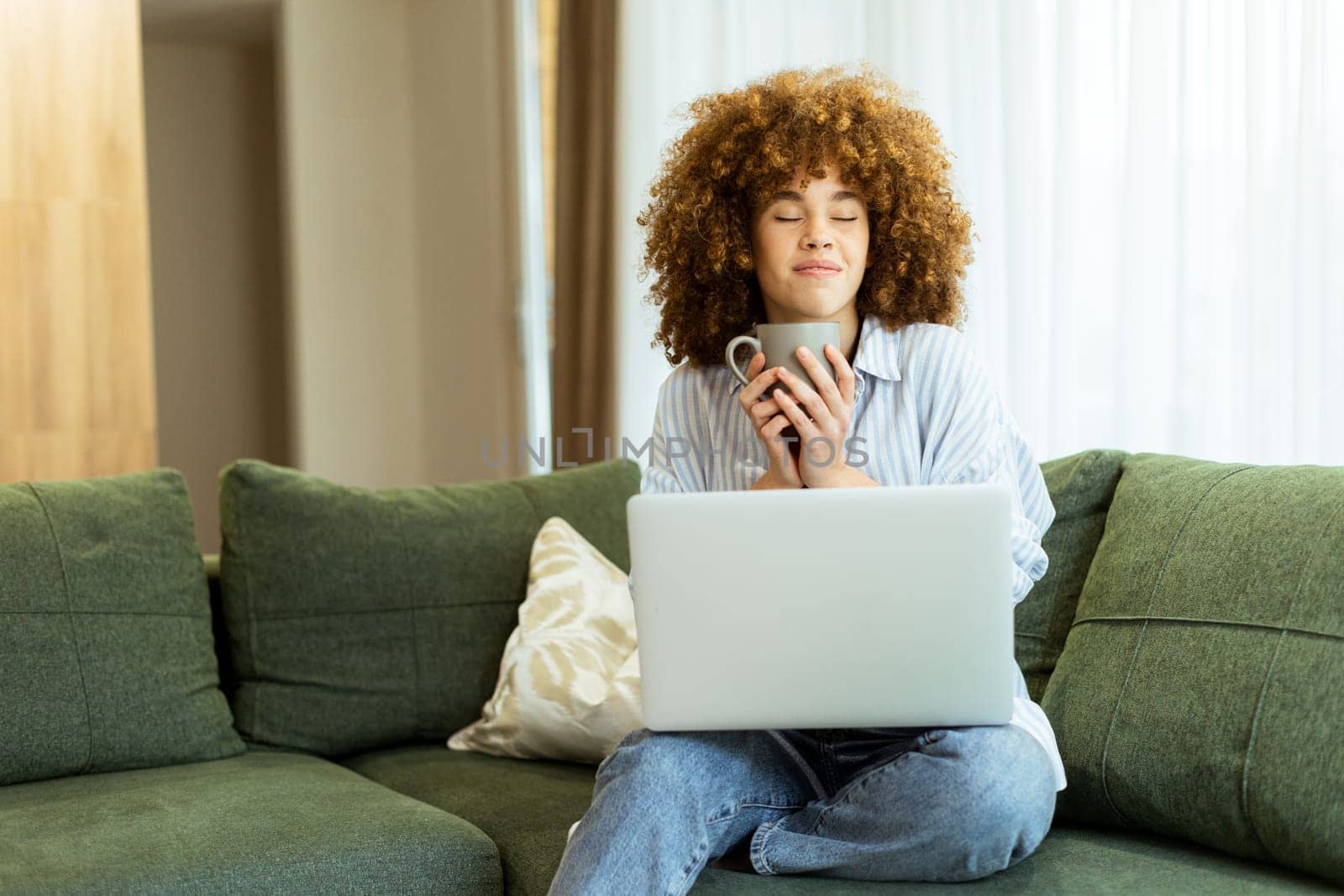 Sitting comfortably on a plush green couch, a young woman savors a steaming cup, embracing tranquility while working on her laptop in a sunlit home environment.