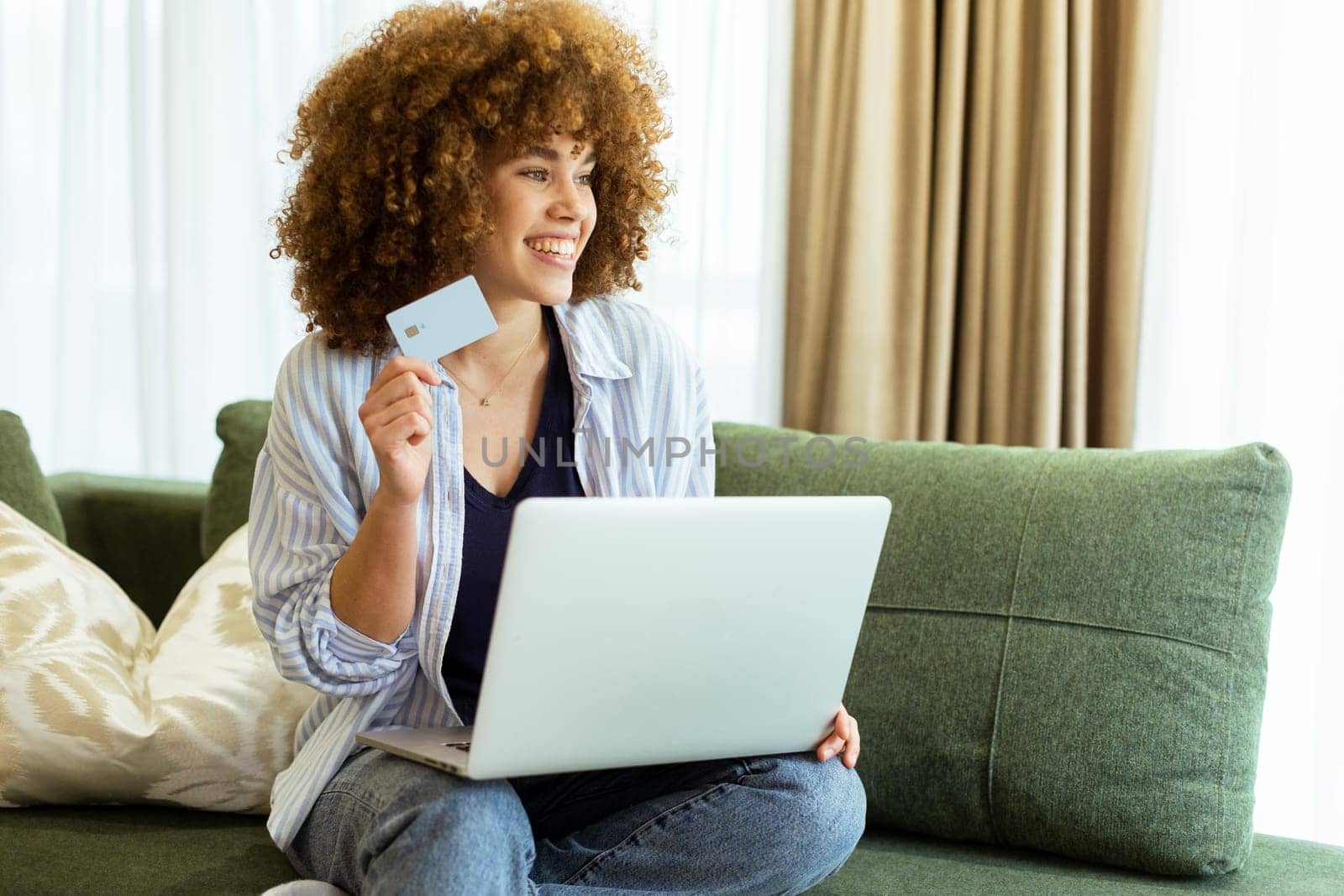 A young woman with curly hair happily shops online, holding a credit card, while seated on a comfortable couch in a stylish living room filled with warm sunlight.