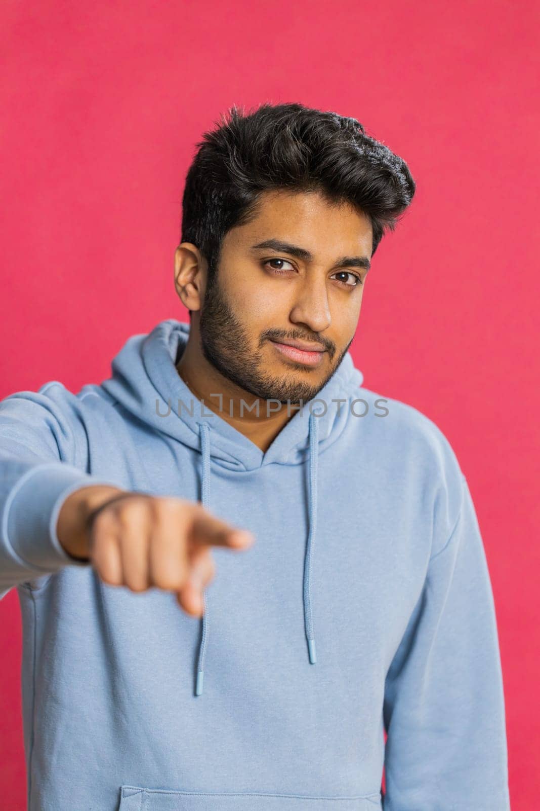 I choose you. Indian smiling man pointing finger to camera and looking with playful happy expression, making winner choice, showing direction. Arabian young guy isolated on red studio background