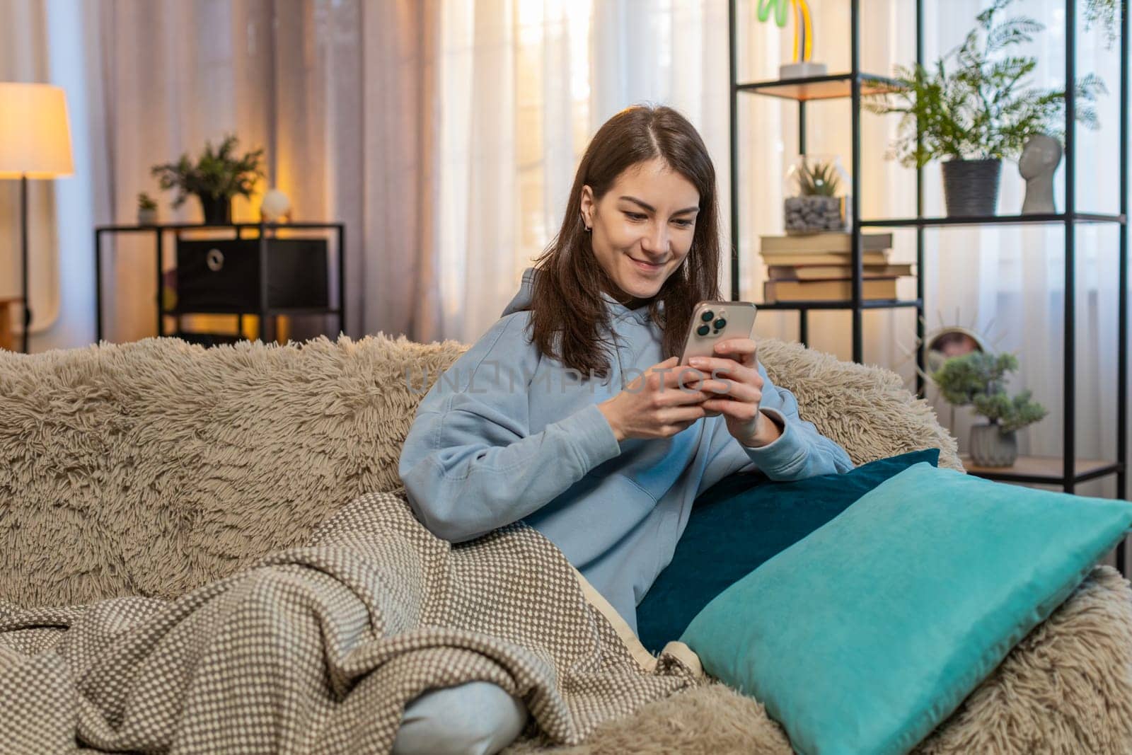 Happy young woman in casual clothes text messaging on smartphone. Caucasian girl covered with blanket lying on couch and using mobile phone in living room at home. Smiling lady relaxing in apartment.