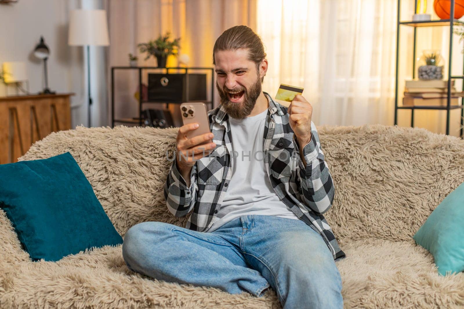 Young man sits at home with phone and credit card, finalizing online store purchase while verifying total. Caucasian guy on sofa presses ok, nodding confidently over successful payment transaction