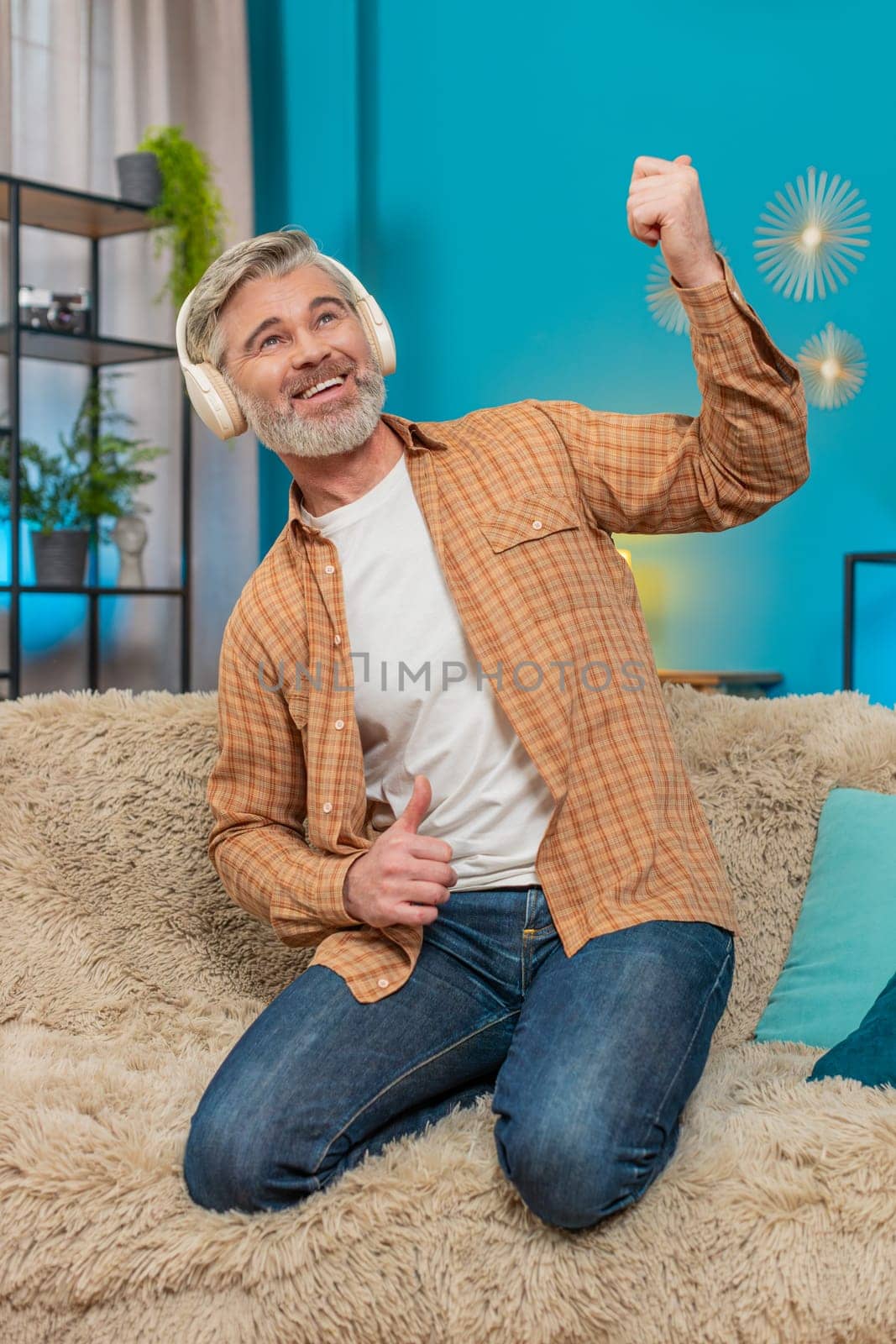 Happy mature man kneeling on sofa at home, wearing wireless headphones. Smiling middle-aged guy energetically dances rock-n-roll, playing an imaginary guitar, enjoying the rhythm of music. Vertical