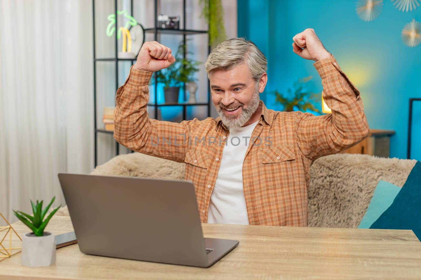 Happy Caucasian mature man using laptop achieve an important email message good news. Satisfied middle-aged guy sitting at home table joyfully exclaims, raising his hands in victory after winning game