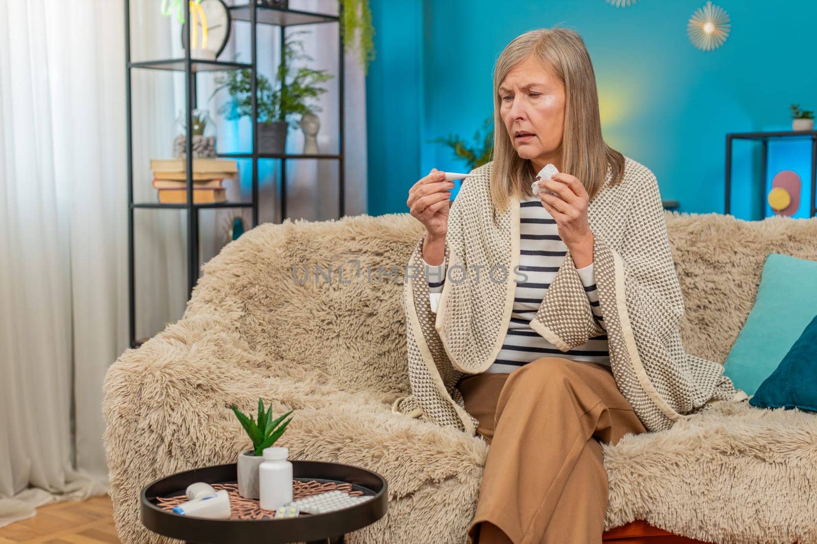 Mature woman sits on sofa covered with blanket checking temperature with thermometer coughing blows sneezes wipes snot into napkin. Grandmother shows signs of illness, including virus allergy symptoms