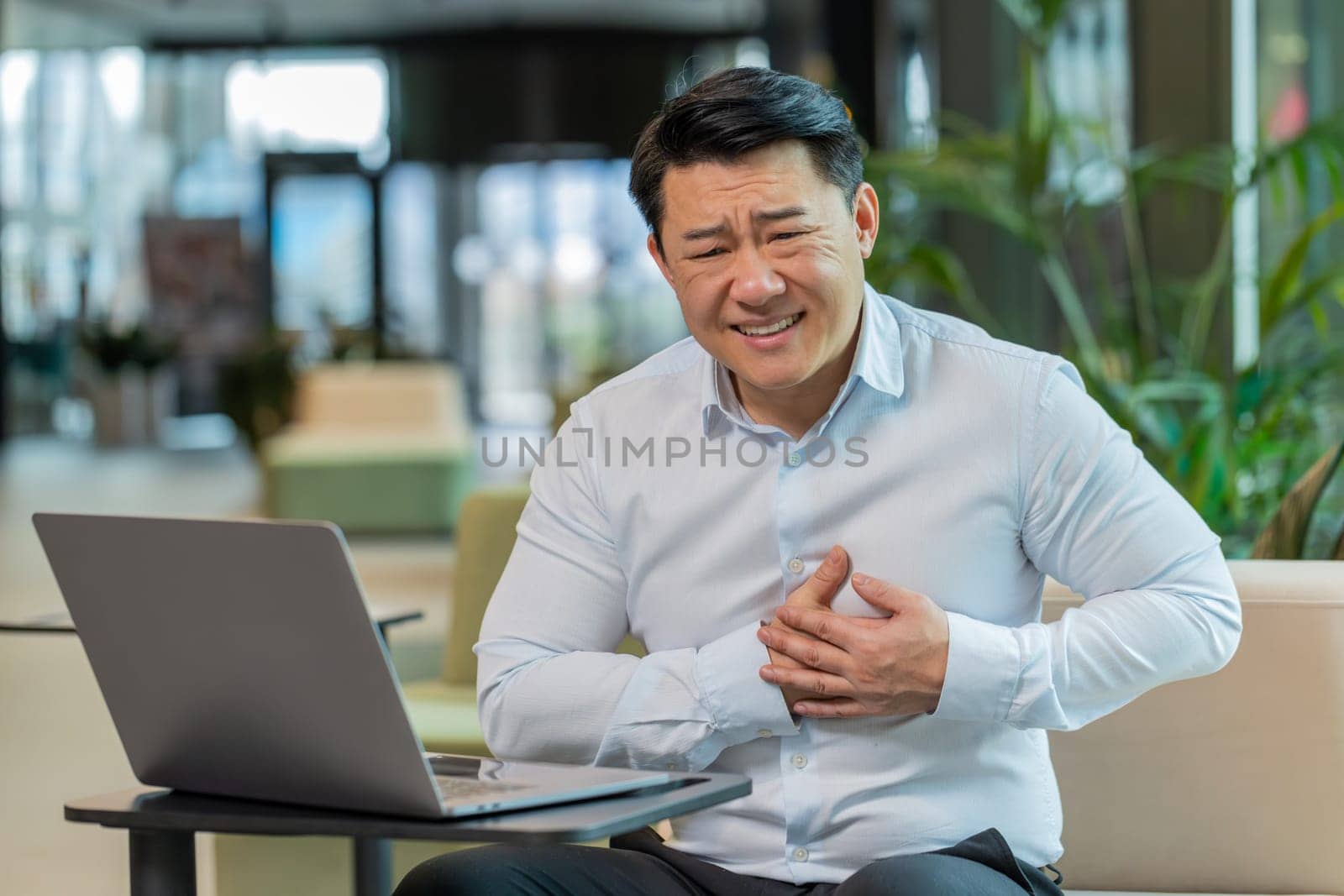 Stressed Chinese mature businessman using laptop and suffers from tension holding chest with heart pain attack risk of stroke symptom while working in modern office lobby. Businessperson in shirt.