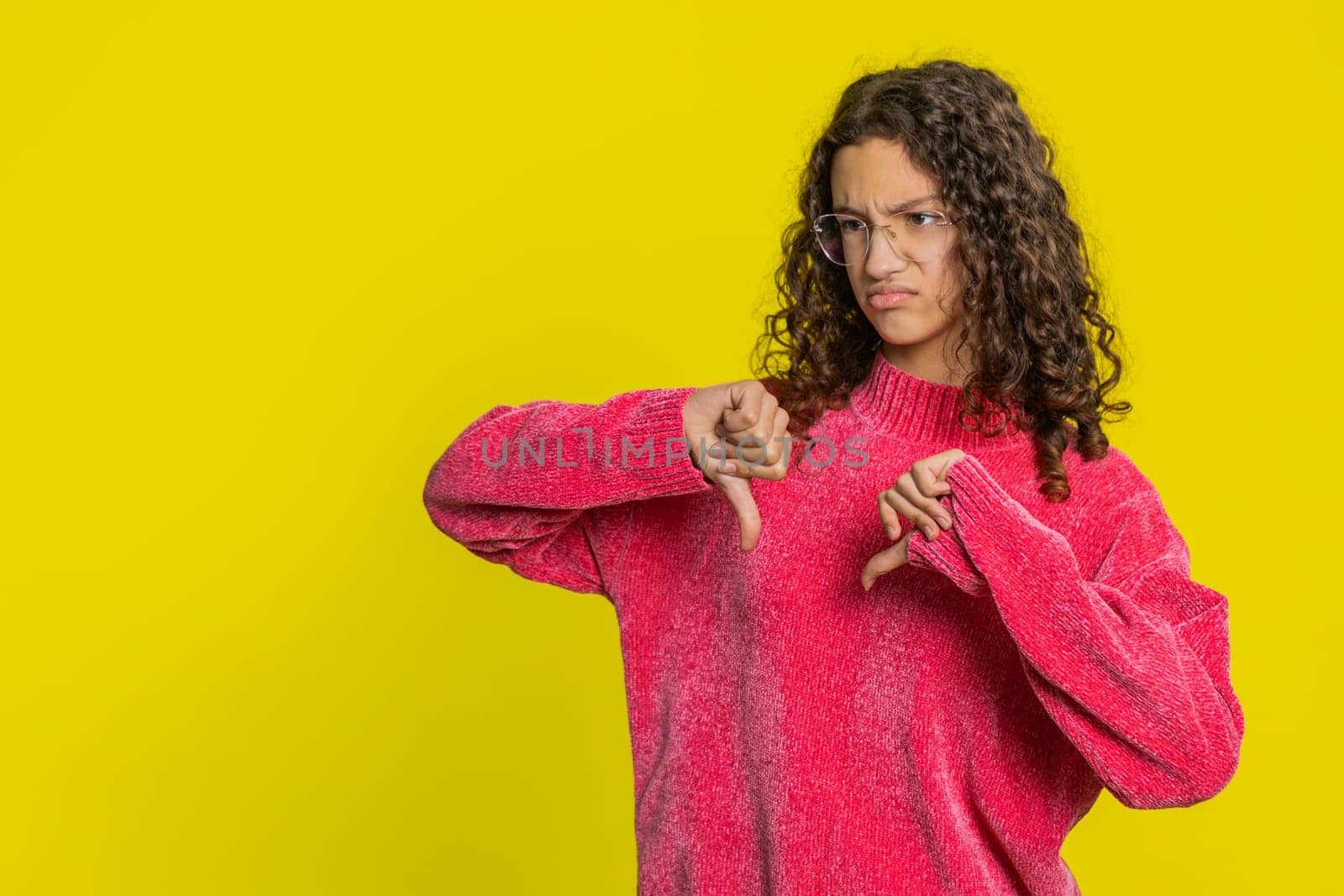 Dislike. Upset unhappy Caucasian girl showing thumbs down sign gesture, expressing discontent disapproval, dissatisfied negative feedback. Dissatisfied teen child indoors isolated on yellow background