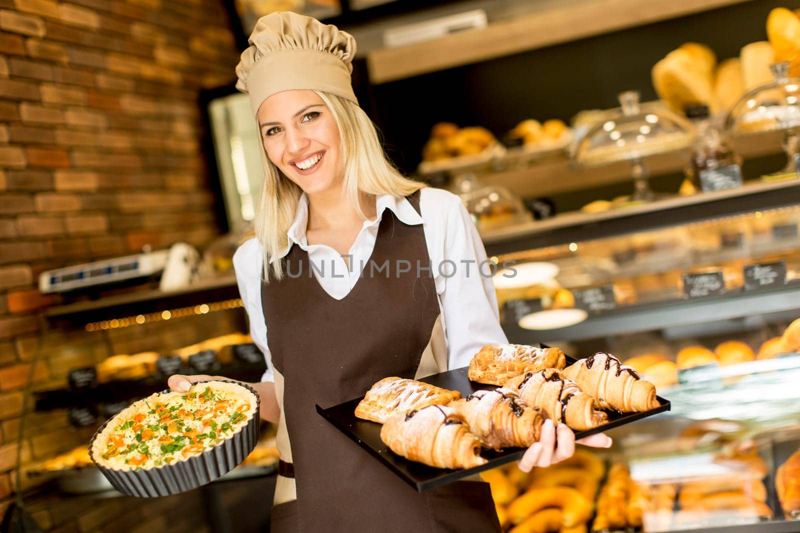 Young female baker standing in the bakery, shows pizza and croissants