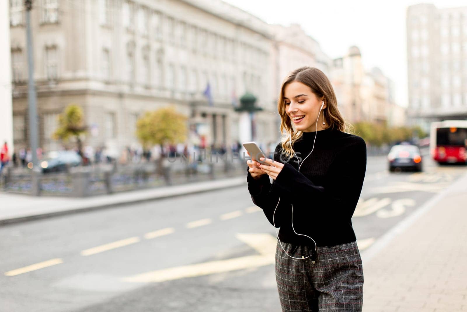 Smiling brunette woman using mobile while standing on street and  waiting for a bus or taxi