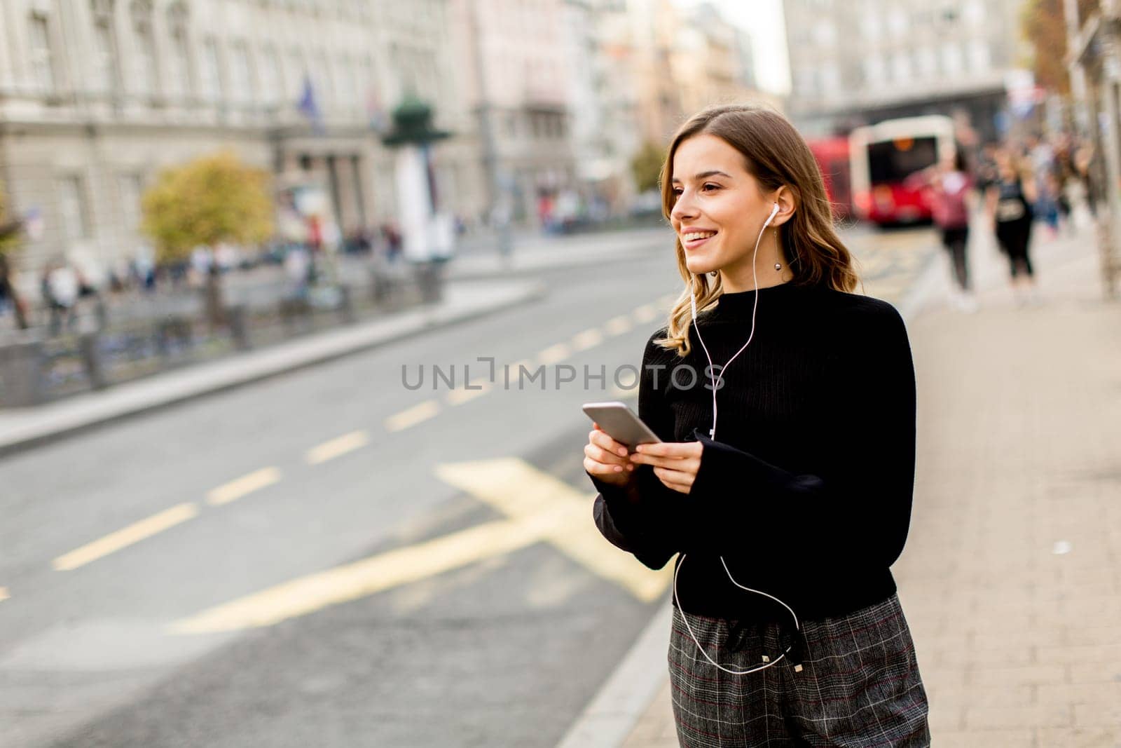 Smiling brunette woman using mobile while standing on street and  waiting for a bus or taxi