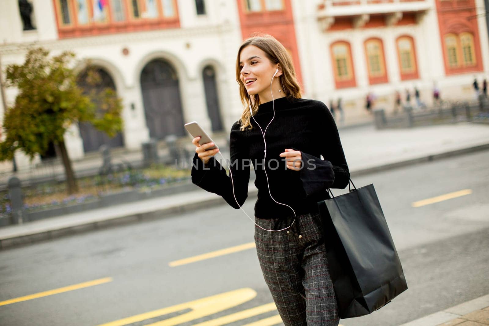 Smiling brunette woman using mobile while standing on street and  waiting for a bus or taxi