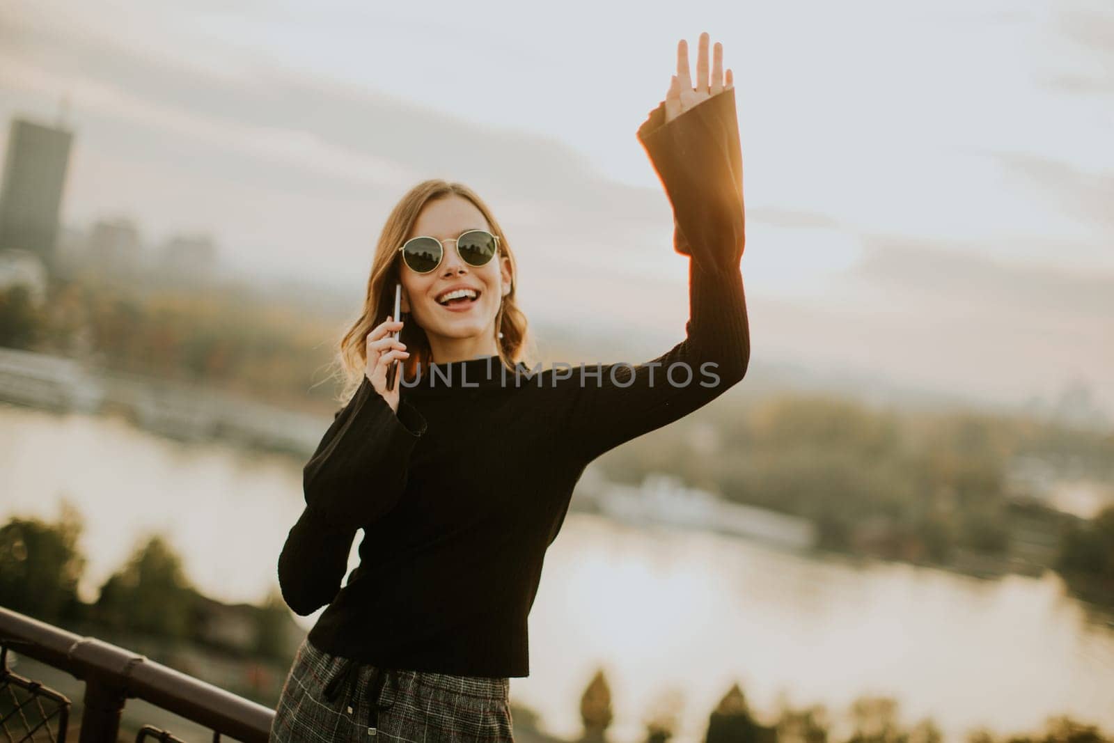 Portrait of young woman holding mobile phone while standing on walkway by the river