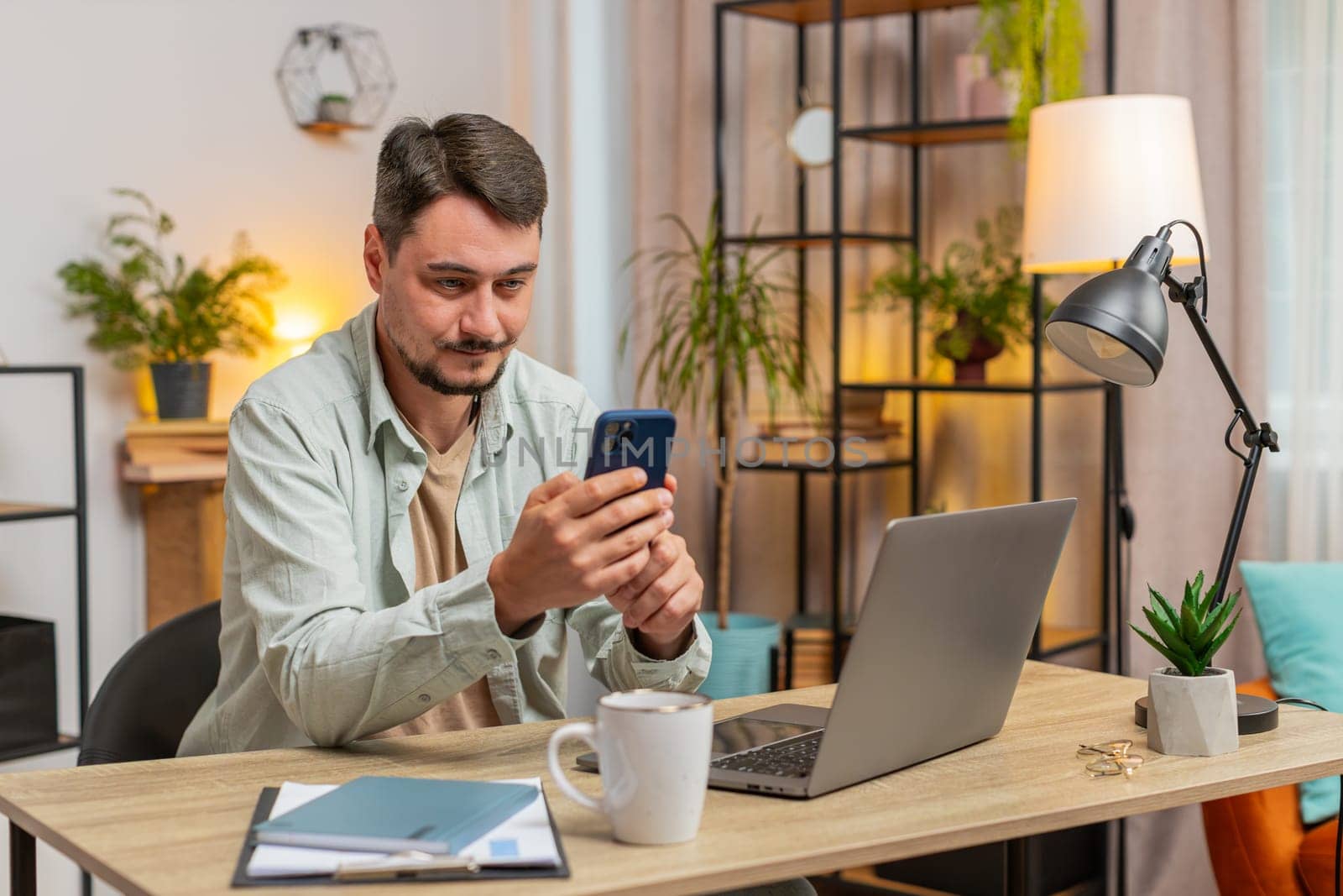 Happy young Caucasian man sitting at table using mobile phone smiling at home office. Guy freelancer texting share messages on smartphone social media applications online watching movie. Lifestyle.