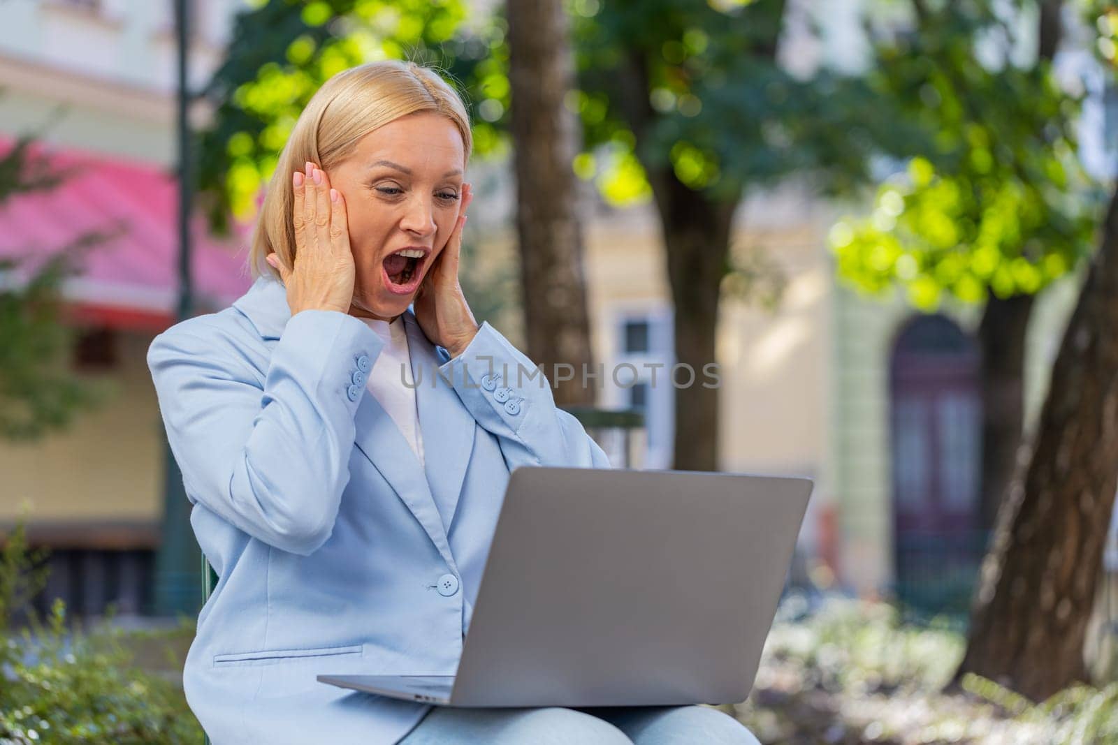 Excited Caucasian mature businesswoman reading great news on laptop computer screen while sitting on bench on city street. Smiling joyful female employer in formal suit celebrating success in business