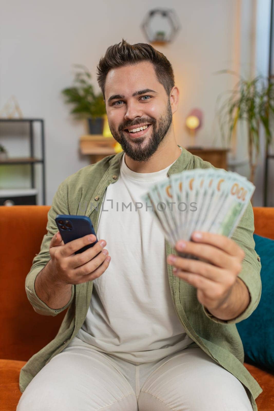 Successful rich young Caucasian man holding and waving dollar bill money fan sitting on sofa couch at home. Happy adult guy winning online lottery game planning vacation calculating budget concept.