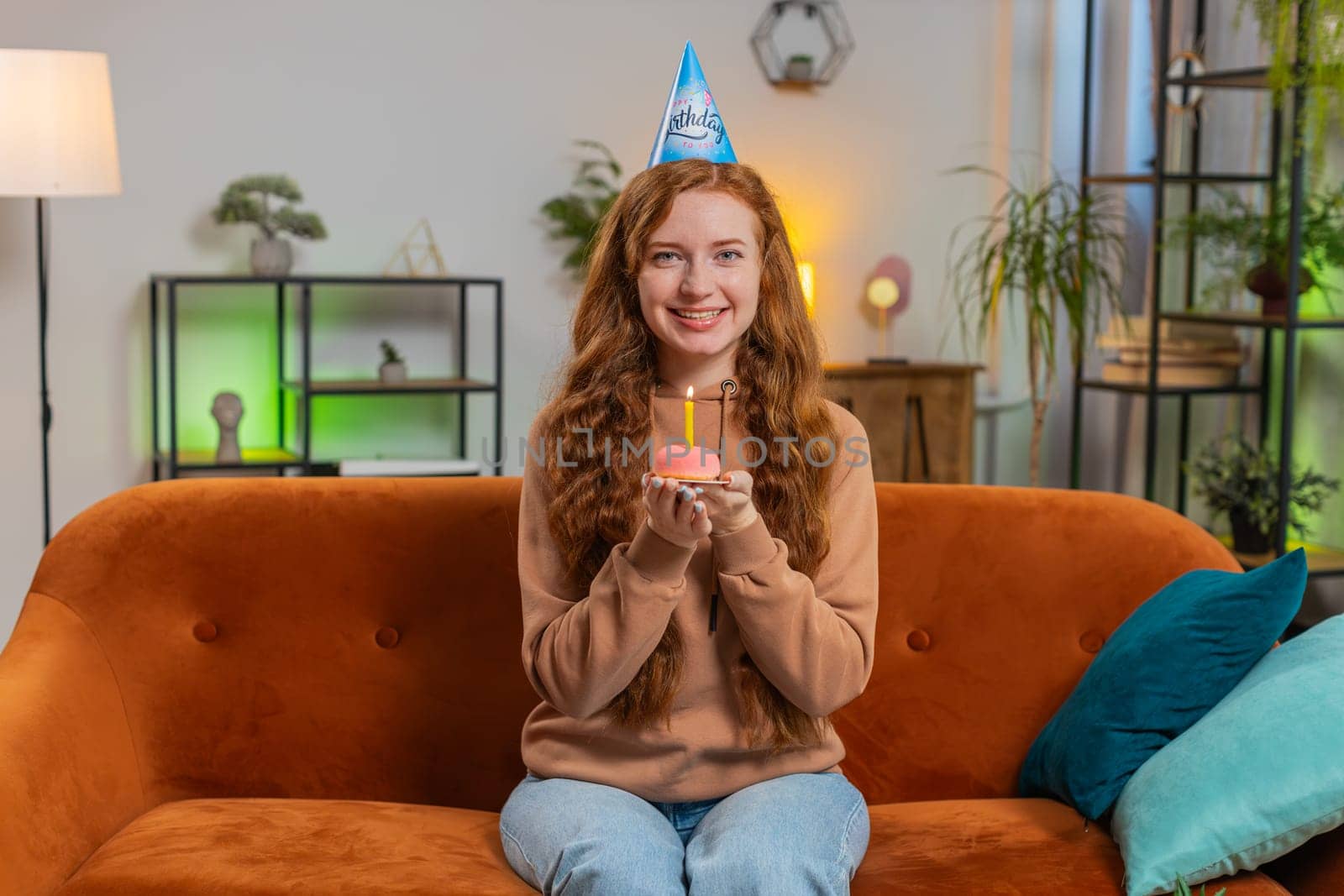 Happy woman wears festive birthday hat holding cupcake makes wish joyful congratulating blowing burning candle on cake. Young redhead girl celebrating birthday party at home room apartment on couch.