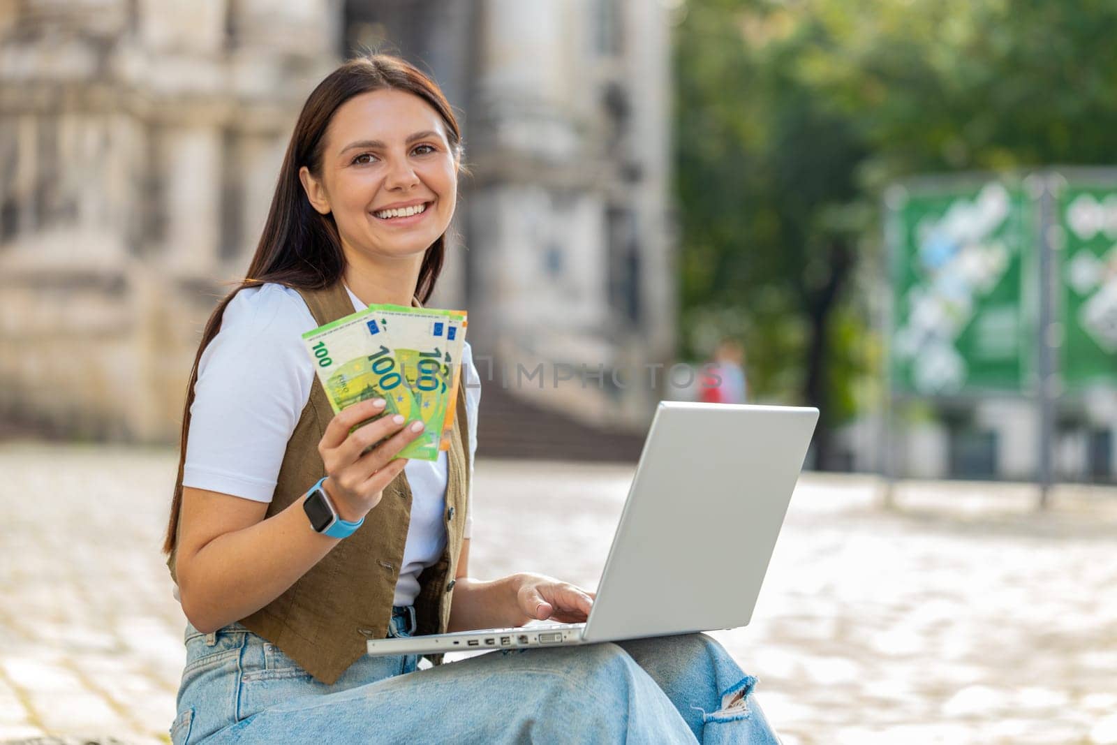 Happy rich woman using laptop computer winner victory holding cash money in euro banknotes sitting on street outdoors during daytime. Young brunette girl tourist planning investment budget success.