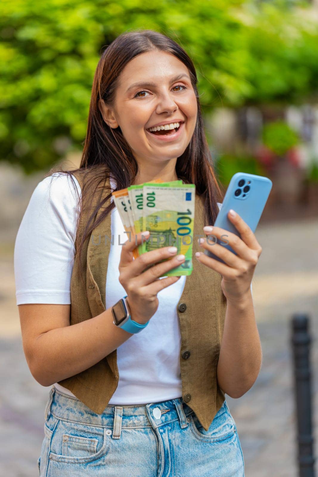 Happy young woman counting money euro cash, using smartphone calculator app, satisfied of income for planned vacation gifts. Lovely girl standing in urban city sunny street. Town lifestyles outdoors