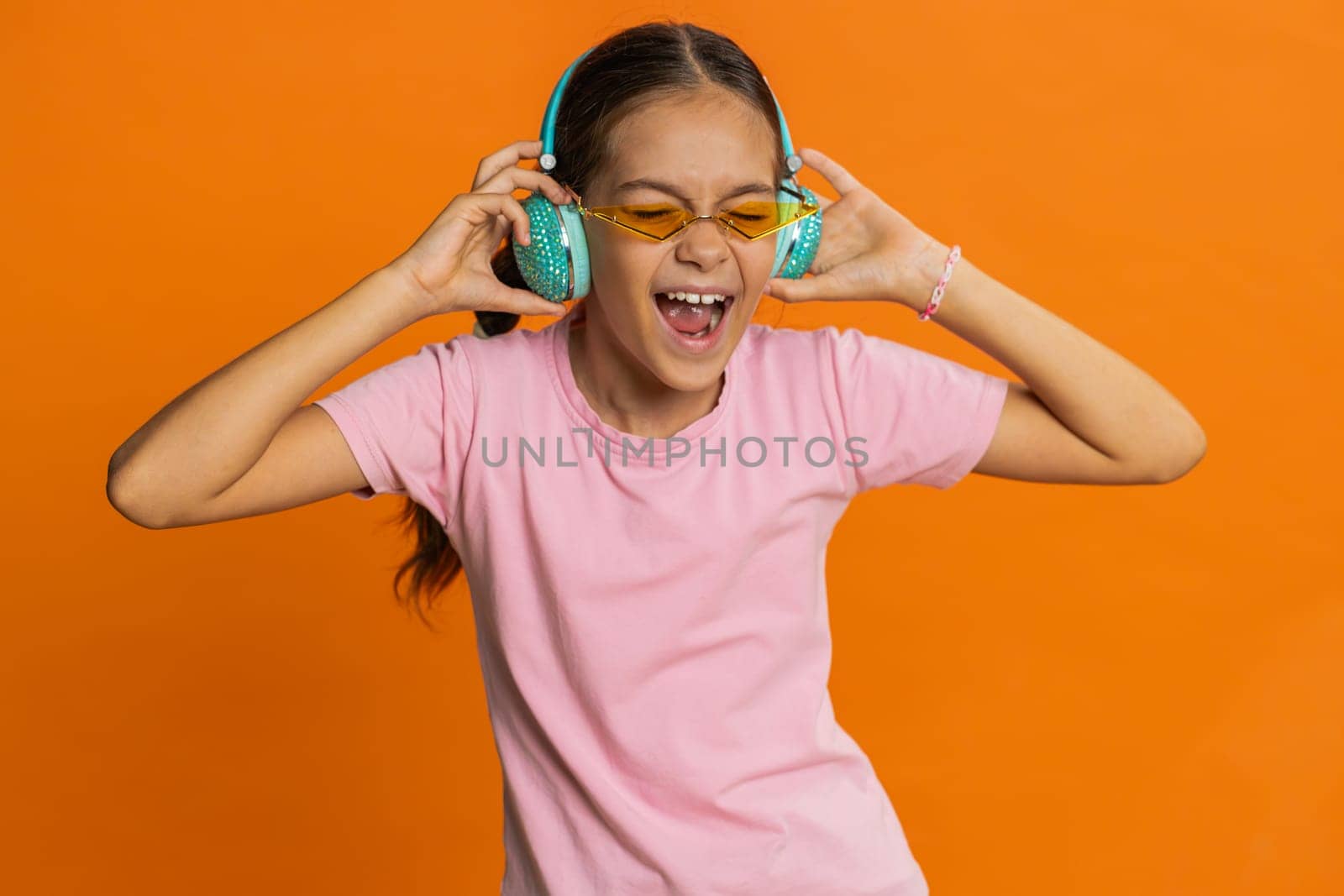 Happy smiling preteen child girl in headphones listening music dancing disco fooling around having fun expressive gesticulating hands relaxing on party making funny moves. Kid on orange background