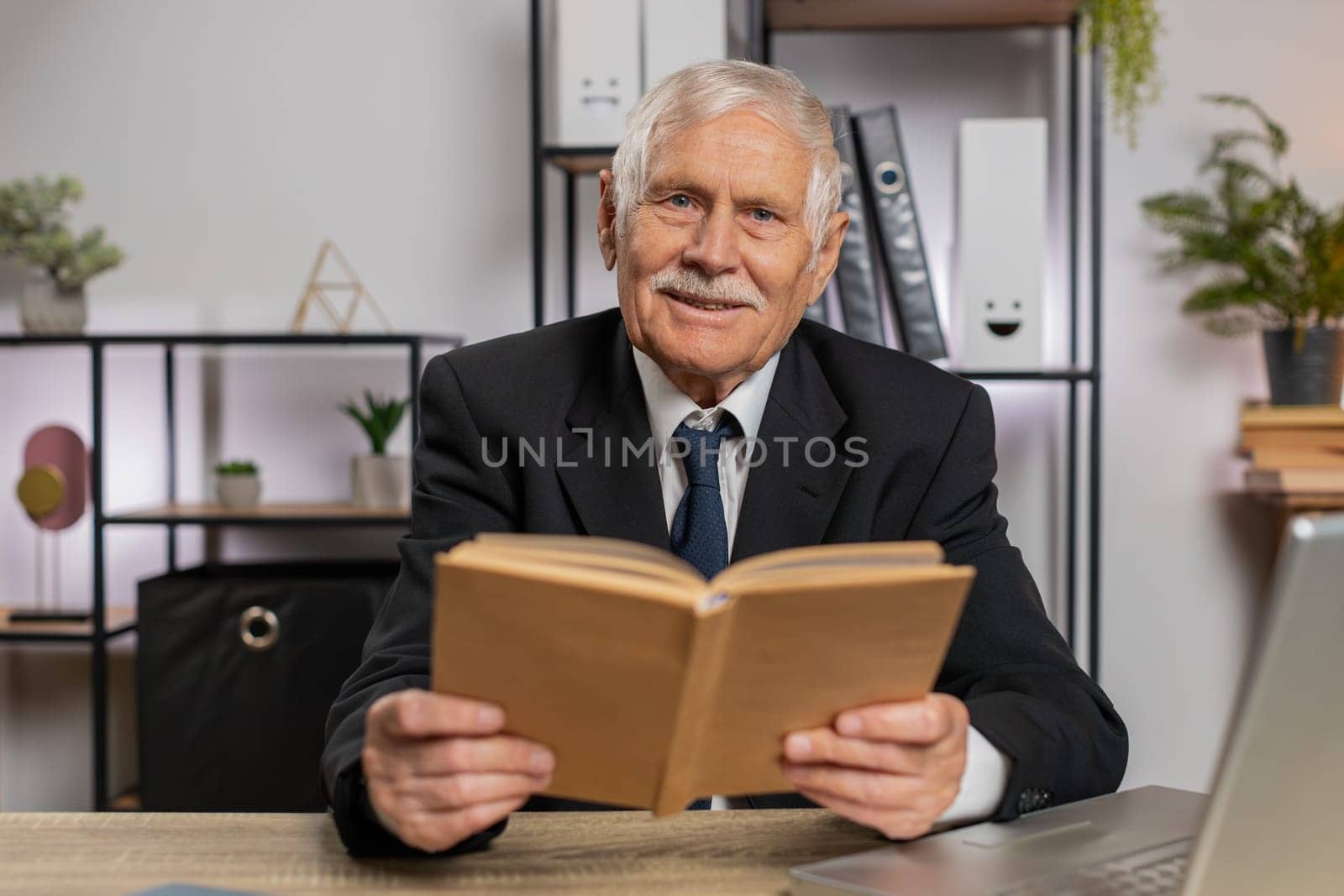 Bored Caucasian senior businessman reading book, literature, taking a rest after work. Grandfather freelancer guy in suit with laptop relaxing with head in hand at home office table. Business people.
