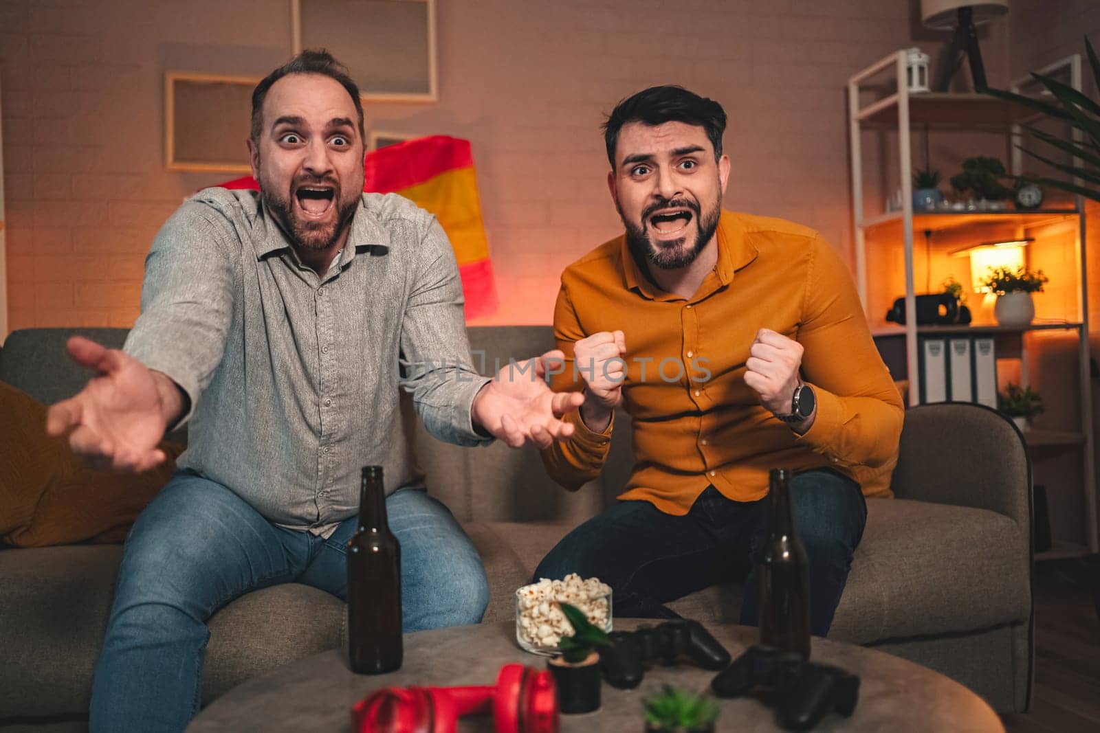 Two friends are wrapped in the thrill of competitive gaming, passionately reacting while seated on a comfy couch surrounded by snacks and drinks in a warmly lit room.