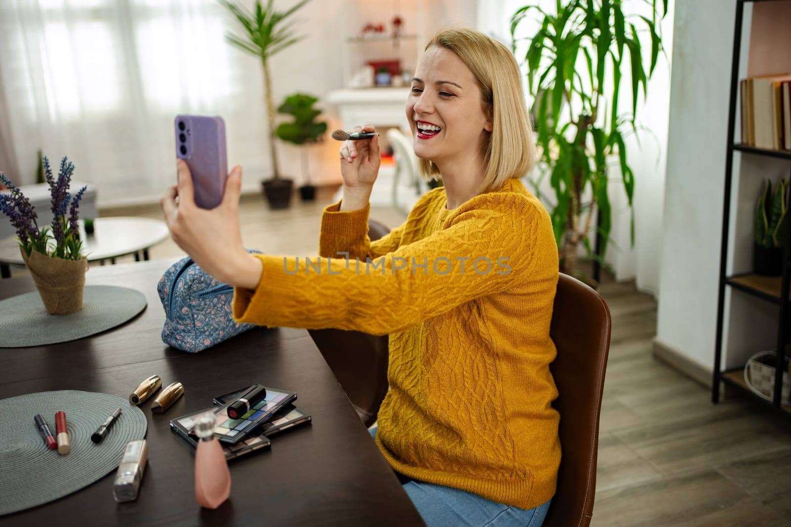 A cheerful woman is engaged in a lively video call, applying her makeup with a mirror in a warmly decorated living space filled with plants. Sunlight streams in, creating a bright atmosphere.