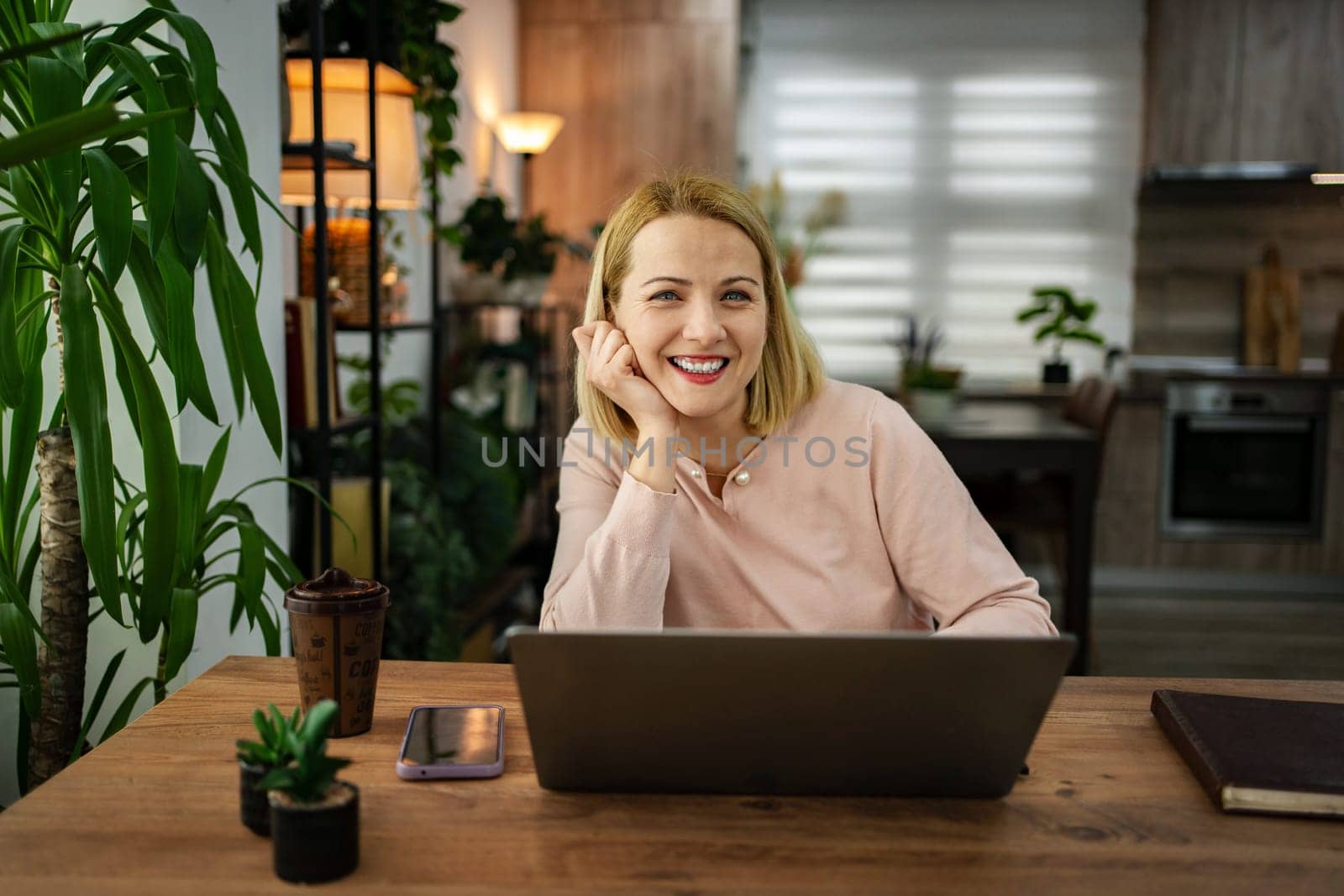 In a warm, inviting workspace filled with plants, a smiling woman sits at a wooden desk, focused on her laptop. Sunlight streams through the window, enhancing the cozy atmosphere.