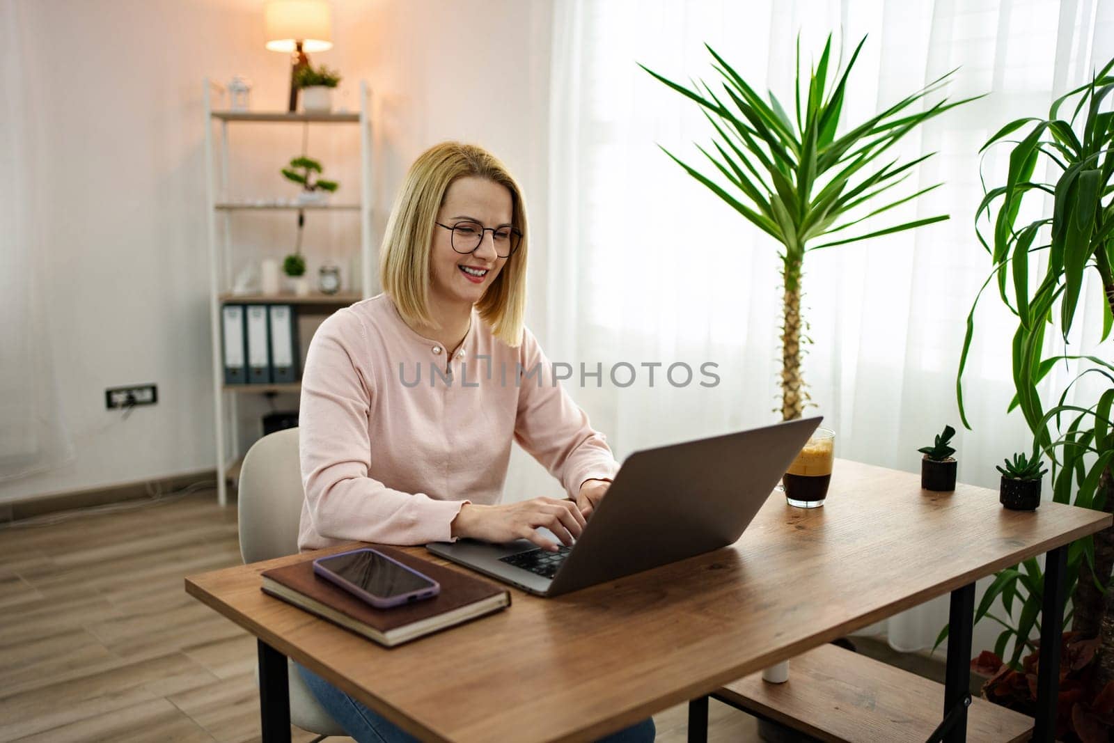A cheerful young woman sits at a wooden desk in a well-lit home office, typing away on her laptop while enjoying a refreshing drink. The space is filled with greenery, creating a tranquil atmosphere.