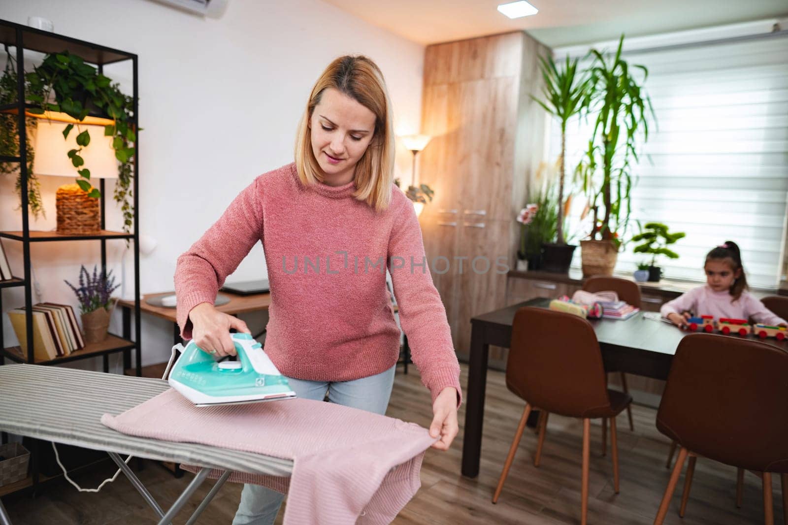 A woman irons clothes while a child plays at a table behind her.
