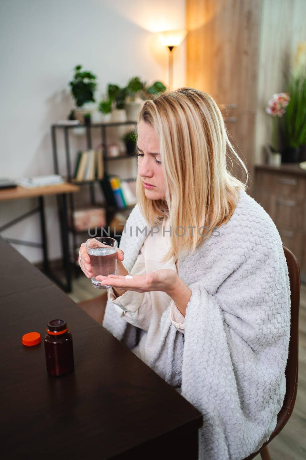 A woman wrapped in a blanket contemplates taking medication in her home.