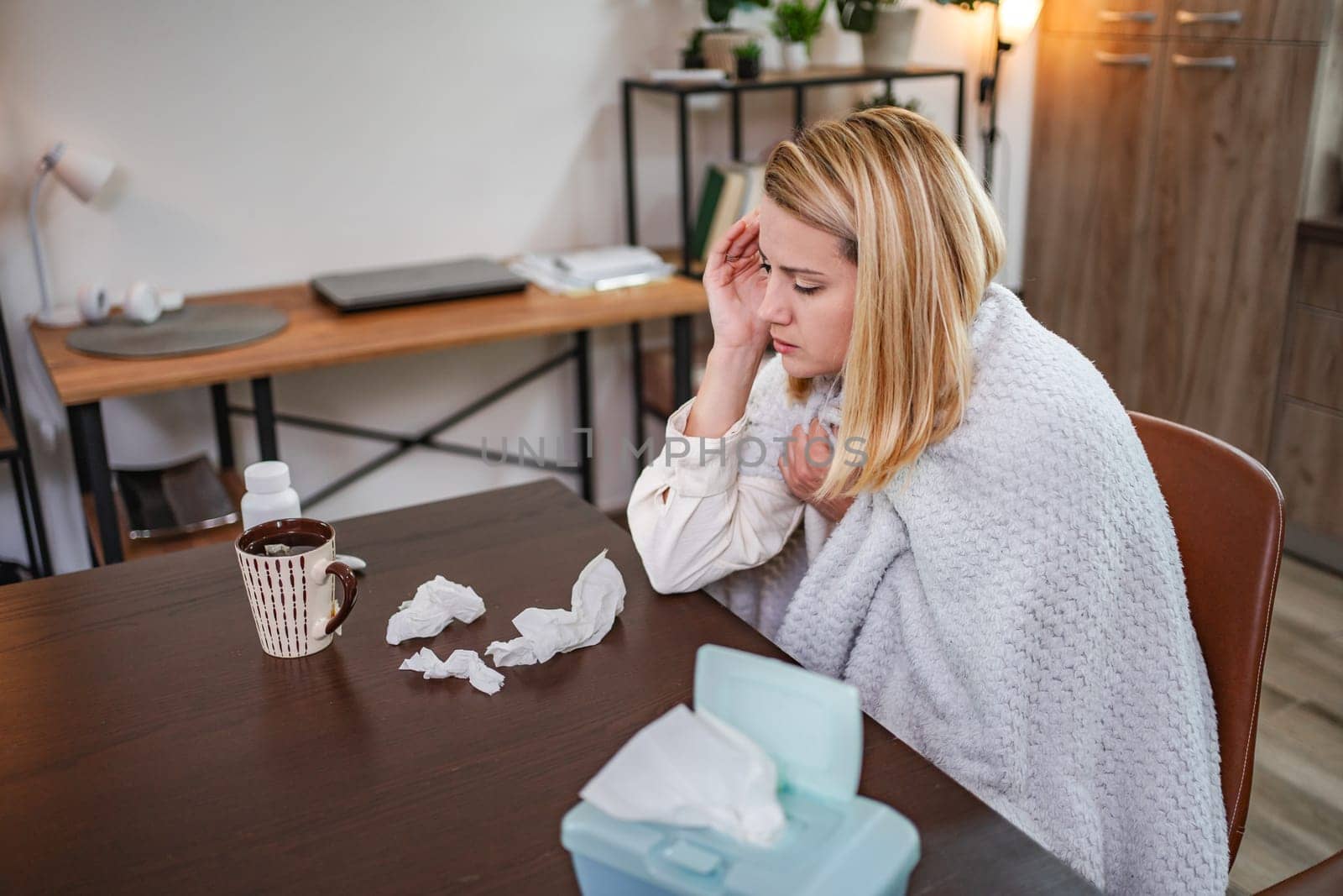 A young woman sits at a table feeling under the weather, surrounded by tissues.