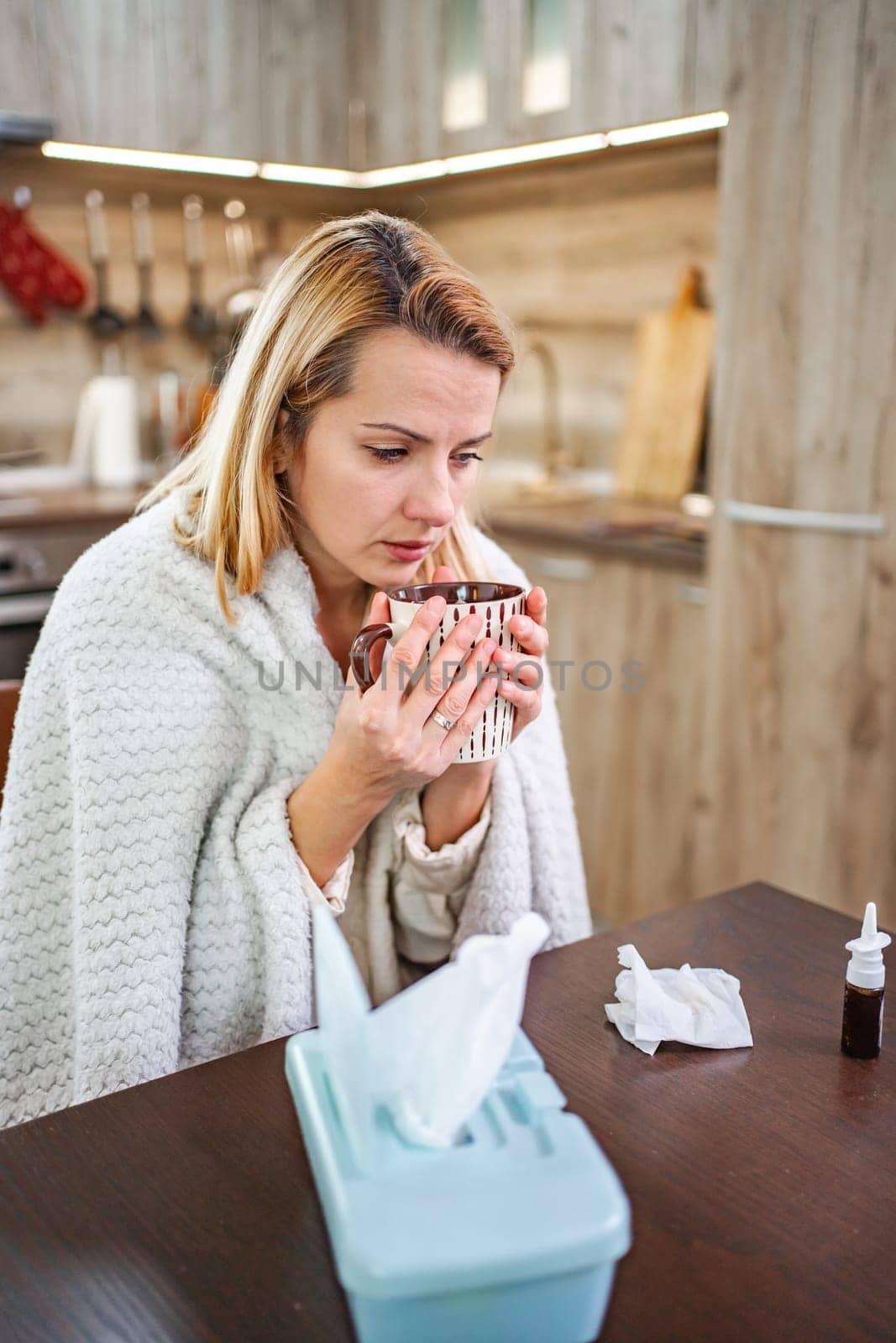 A woman wrapped in a blanket enjoys tea while recovering at home.