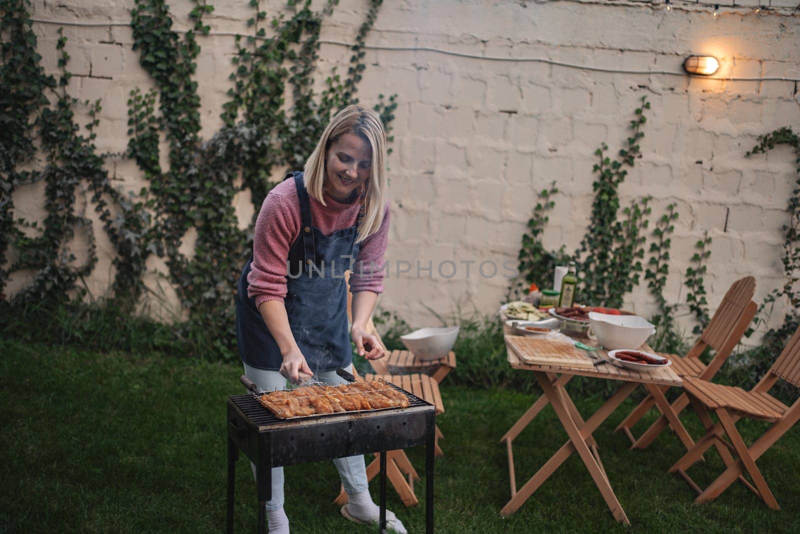 A joyful woman prepares flavorful skewers on a grill in a serene garden space.