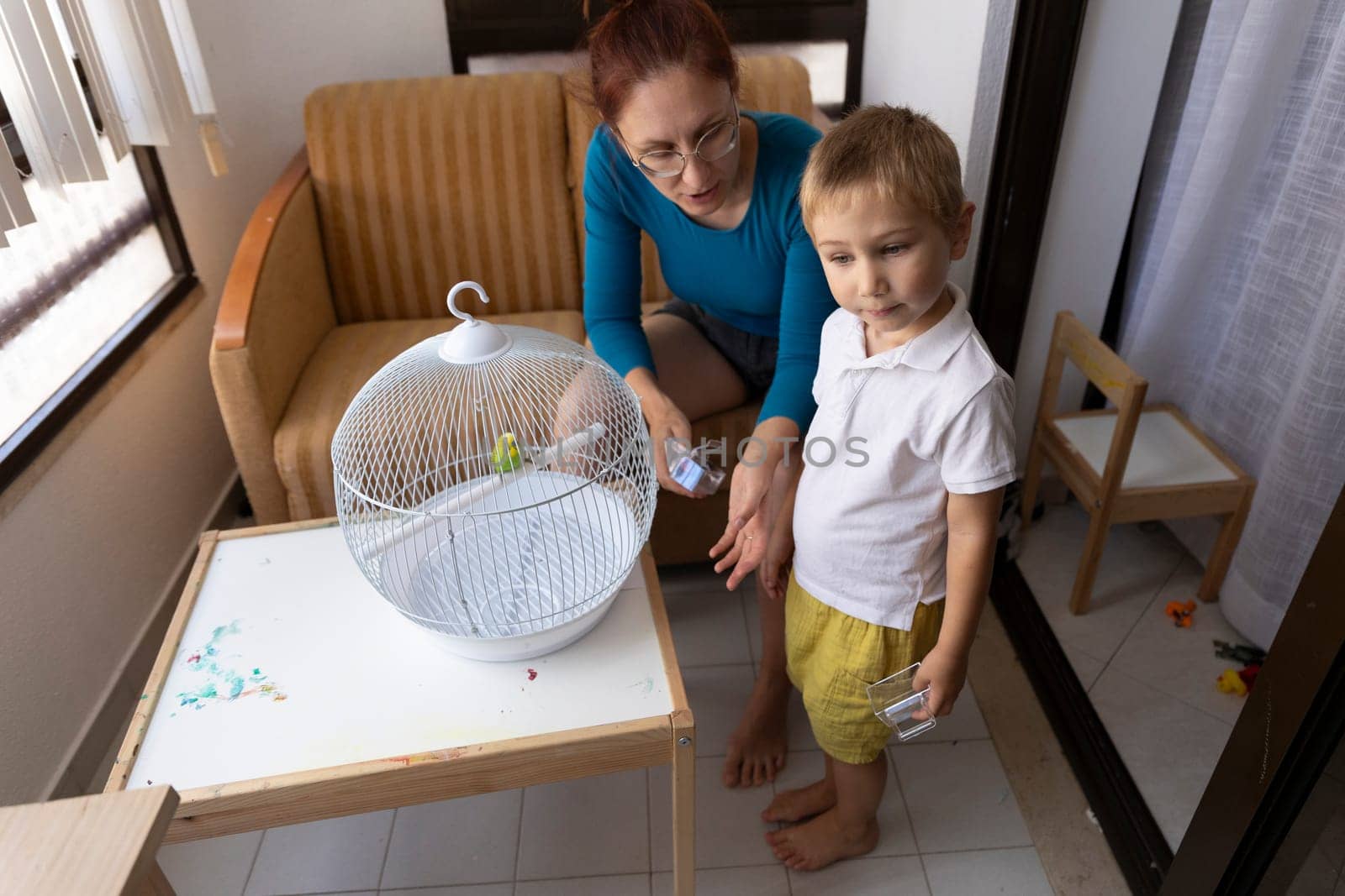 Mother and son interacting with their pet budgie in a birdcage, demonstrating responsible pet ownership and family bonding