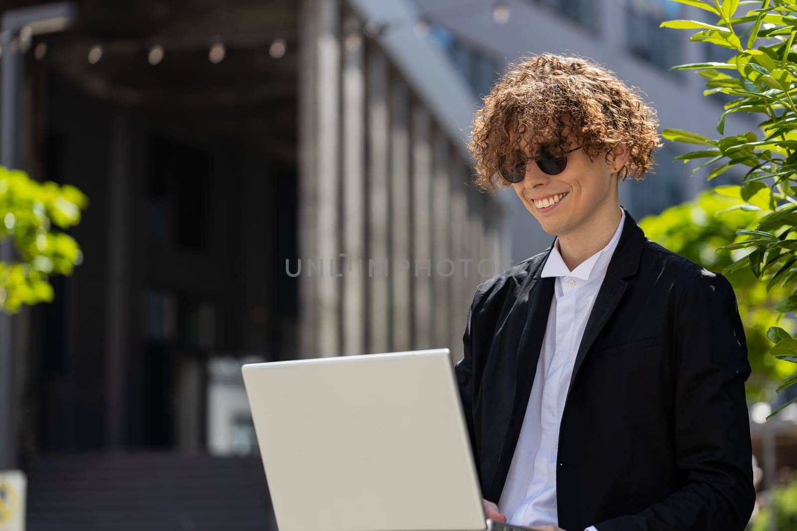 Caucasian young businessman investor employer entrepreneur sitting outside office building opening laptop and working on business project in city. Freelancer guy man in elegant suit and sunglasses.