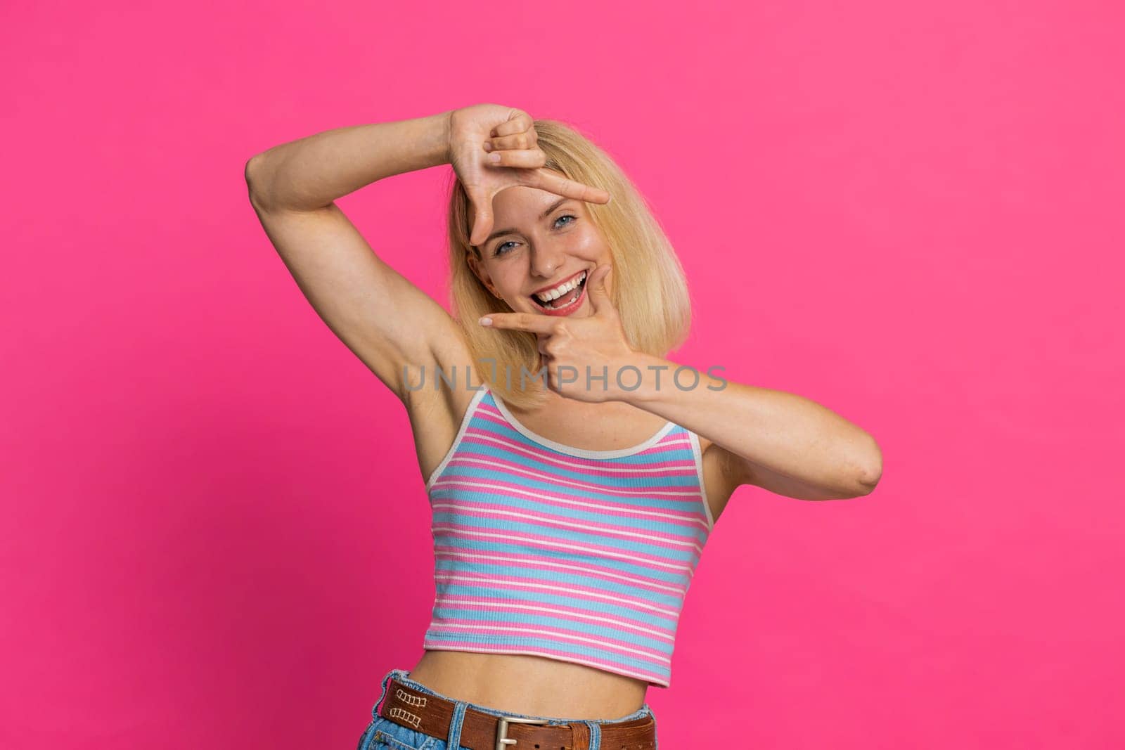 Photographer young blonde woman gesturing picture frame with hands, looks through fingers and focusing on interesting moment imitating zoom and cropping nice image. Girl isolated on pink background
