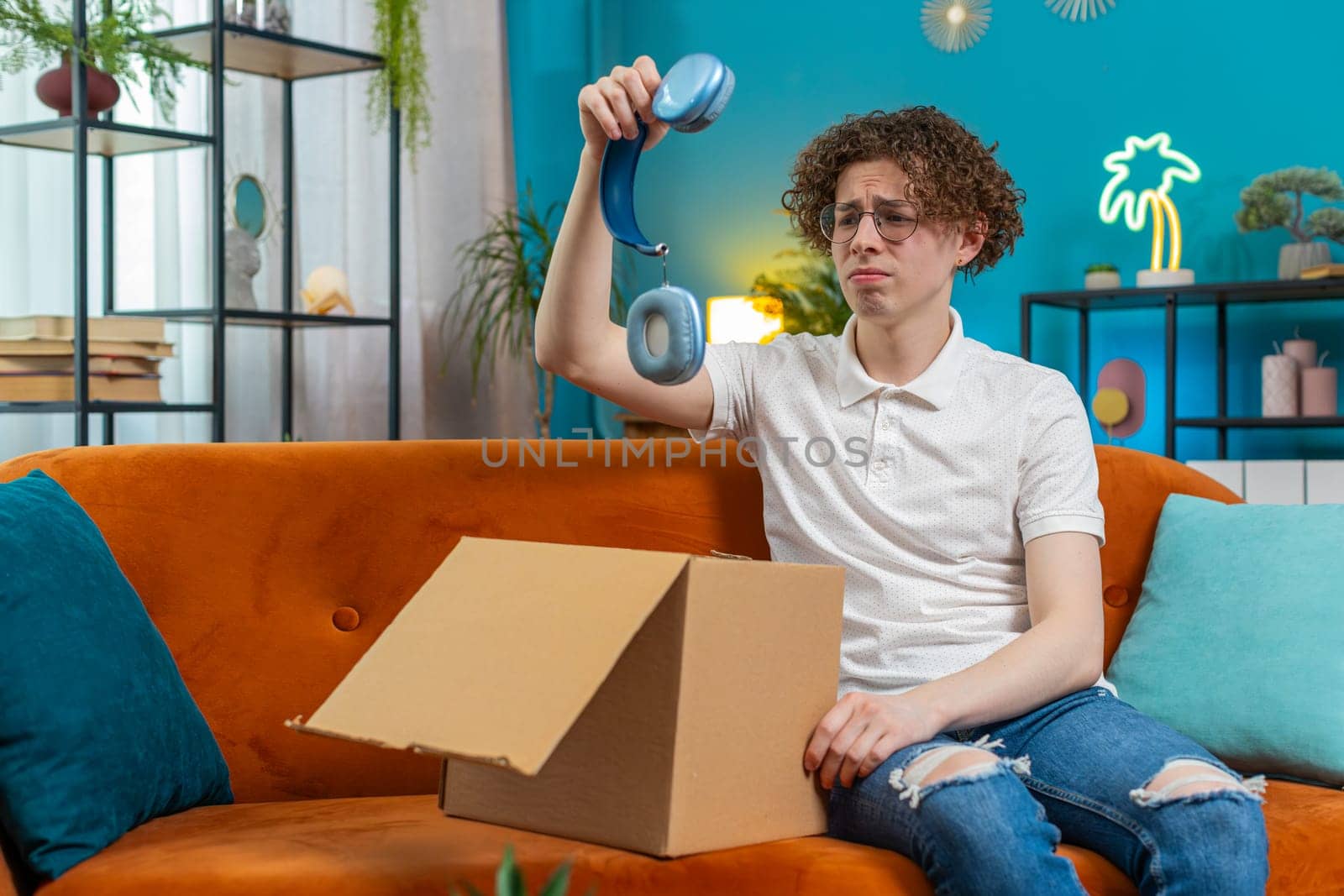 Unhappy young Caucasian man guy unpacking carton box, feeling dissatisfied with getting damaged wireless headphones order, writing negative feedback in mobile application, internet shopping concept.
