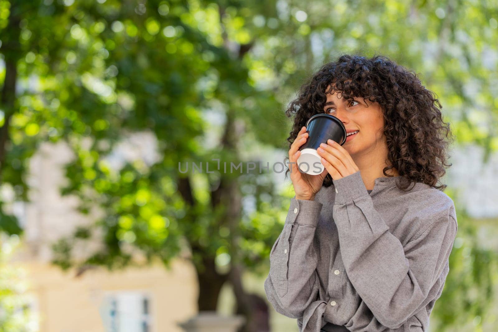 Happy Caucasian young woman enjoying morning coffee hot drink and smiling outdoors. Relaxing, taking a break. Lady girl walking on urban city street drinking coffee to go. Town lifestyles. Sunny park