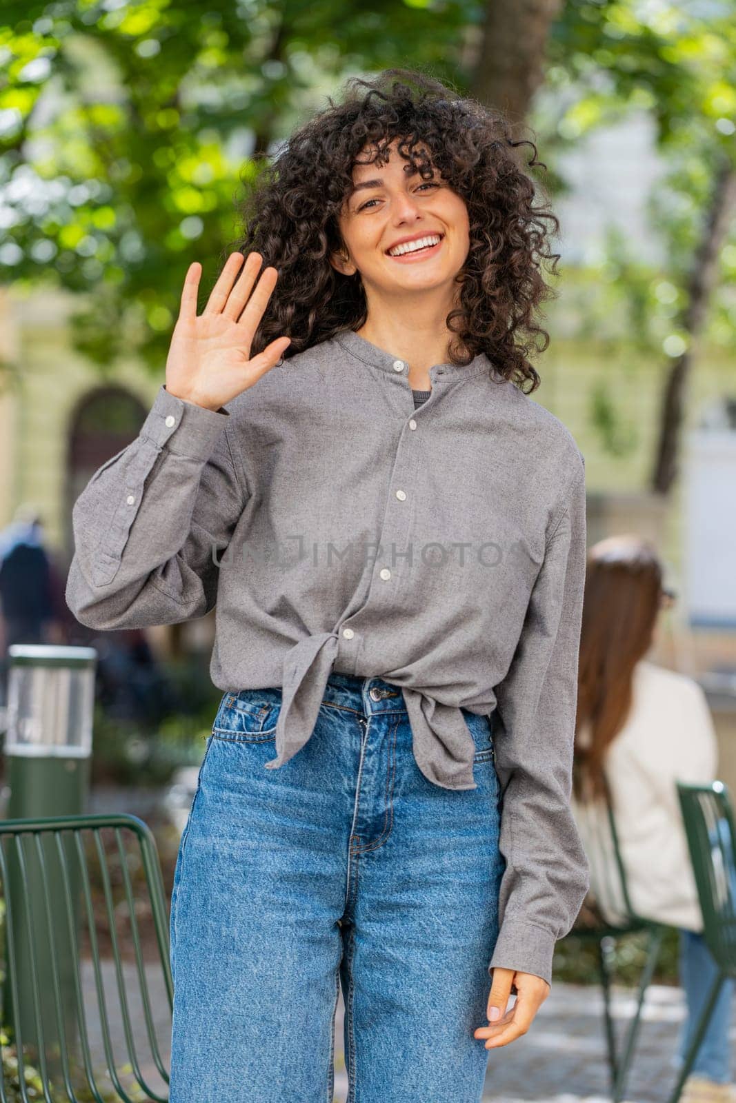 Hello. Caucasian young woman smiling friendly at camera, waving hands gesturing hi, greeting or goodbye, welcoming with hospitable expression outdoors. Lady girl on urban city street. Vertical
