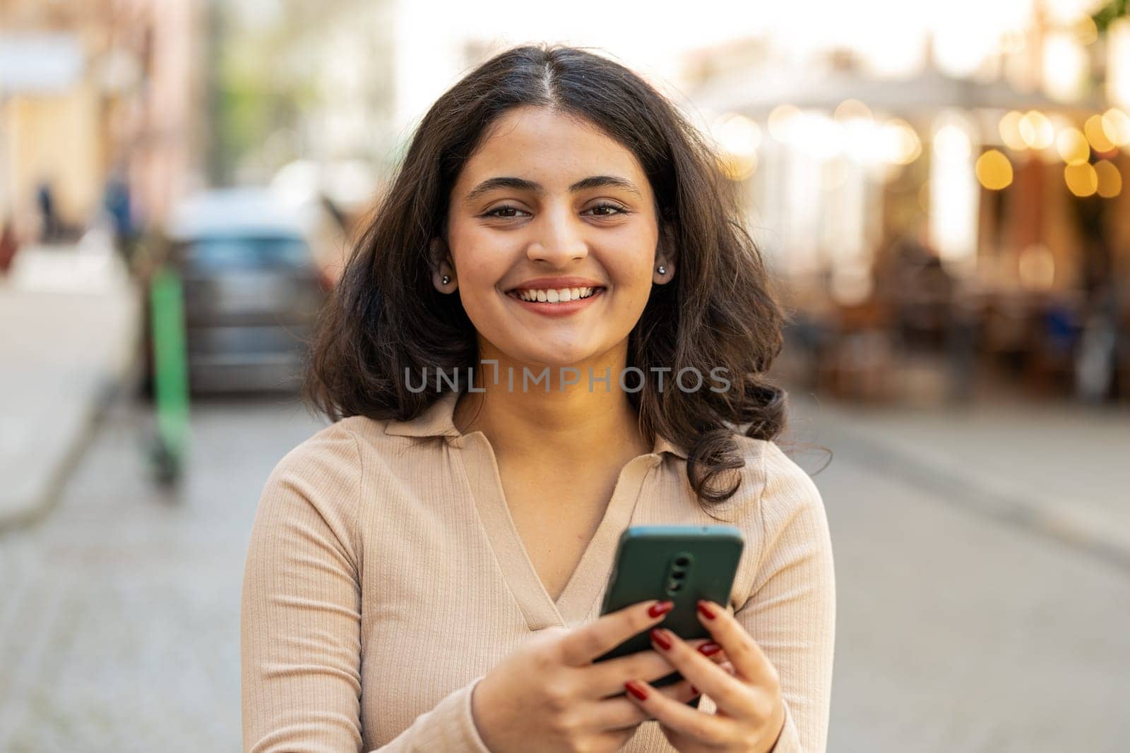 Happy smiling Indian young woman using smartphone typing text messages browsing internet, finishing work, looking at camera outdoors. Hispanic girl walking on urban city street. Town lifestyles