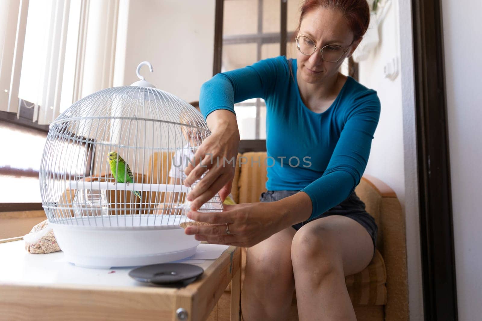 Woman changing water in a white round bird cage with a green parrot perched on a wooden stick