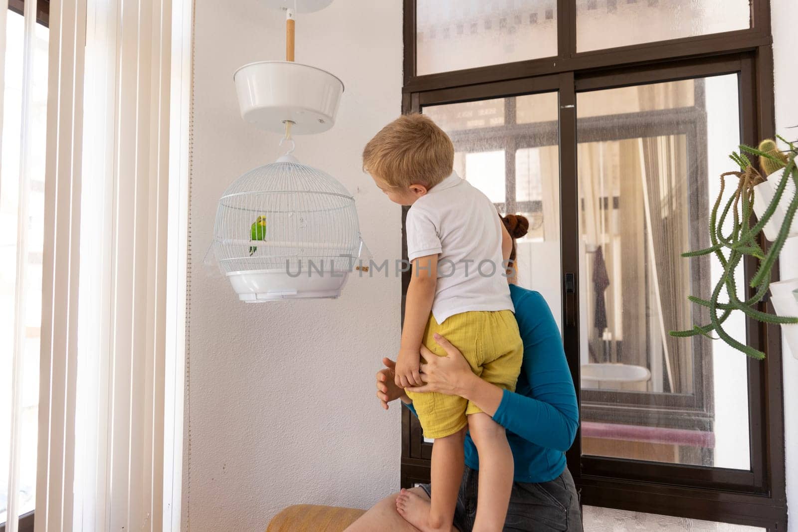 Mother holding her son so he can look at a green parrot in its cage hanging from the ceiling